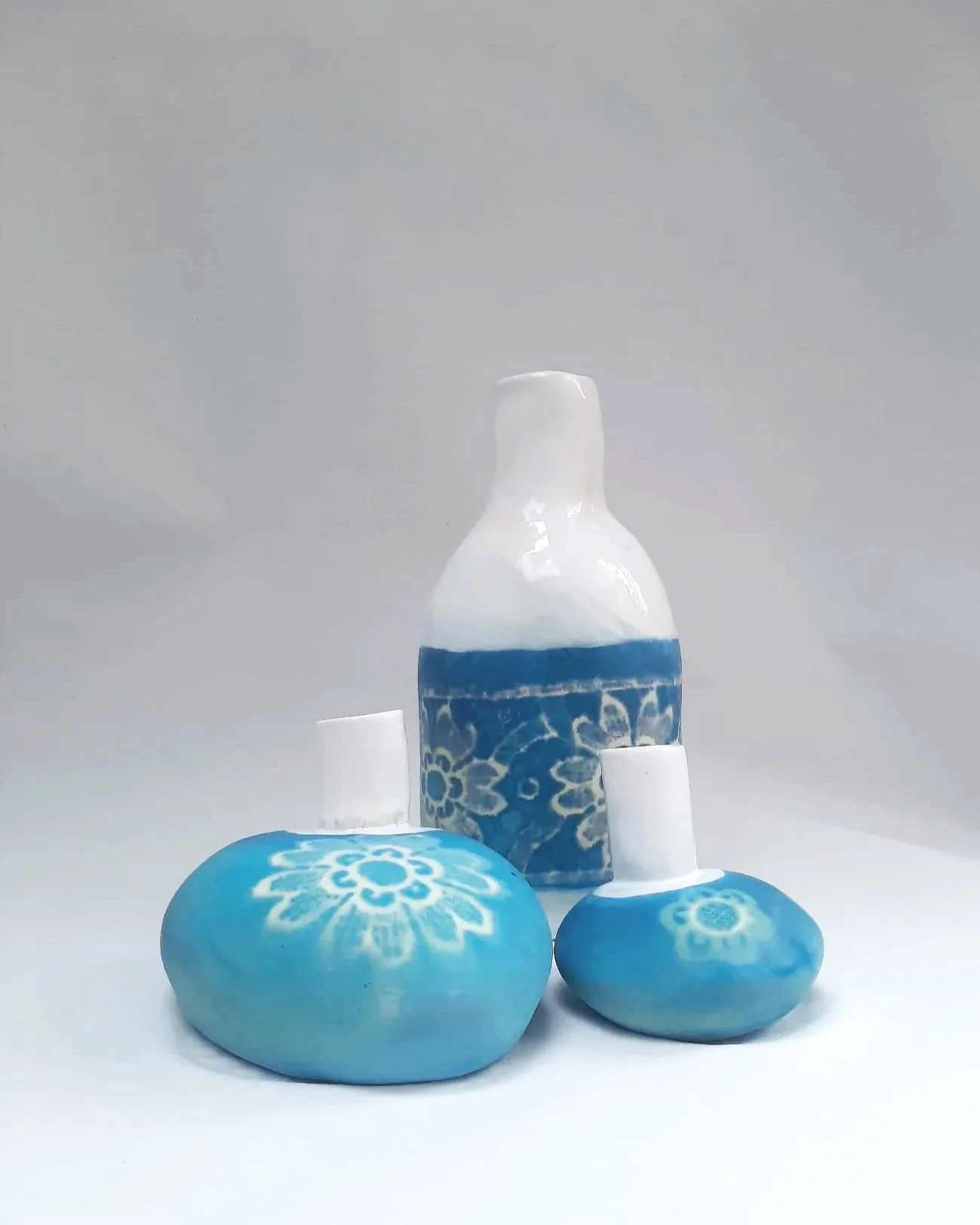 Trois objets en céramique bleue avec des motifs floraux blancs, un vase et deux petits bocaux, sont placés sur une surface blanche contre un fond neutre.