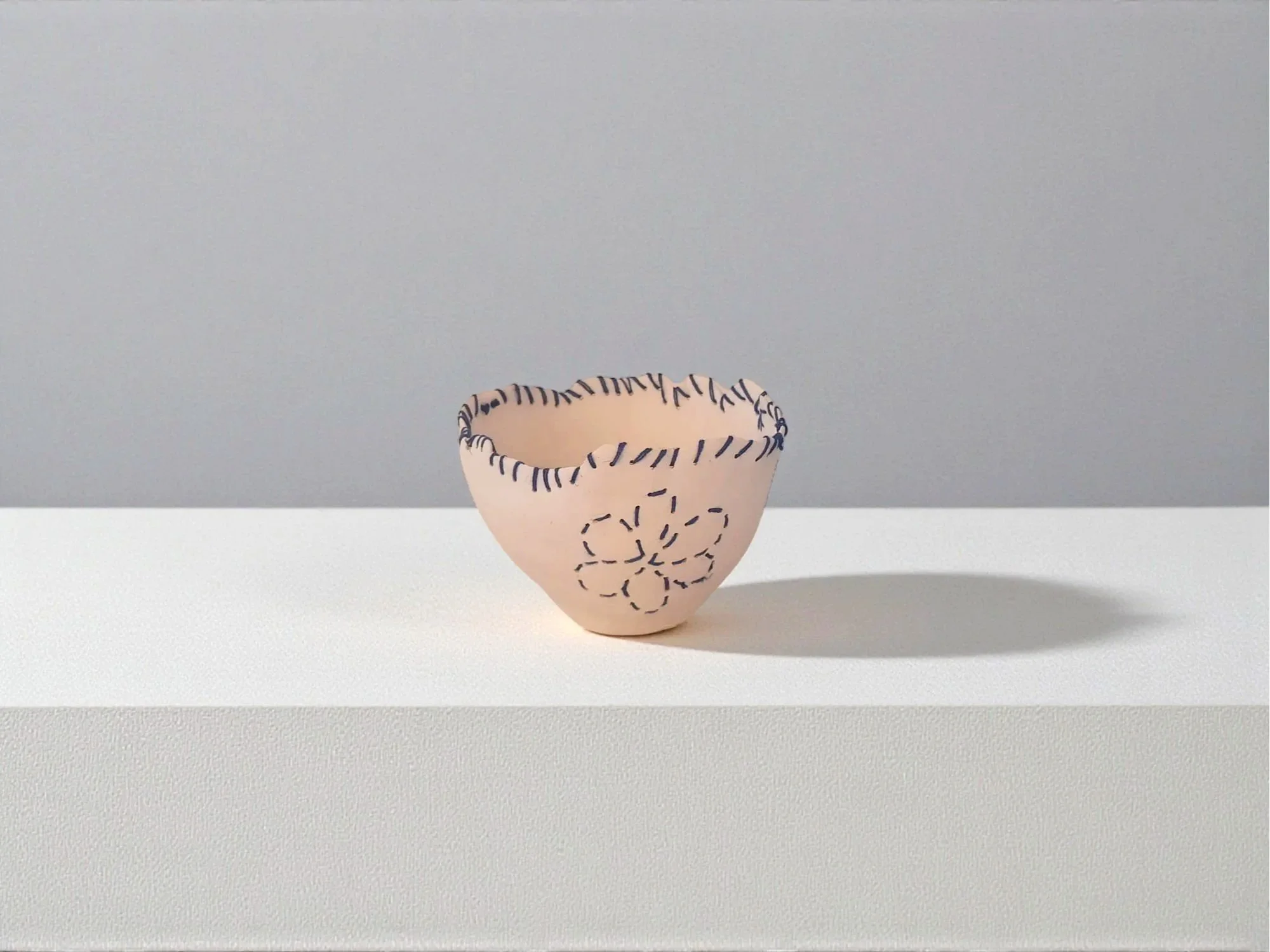 Un bol en céramique rose clair décoré de lignes noires et d'une fleur stylisée sur le devant, placé sur une surface blanche avec un fond gris clair.