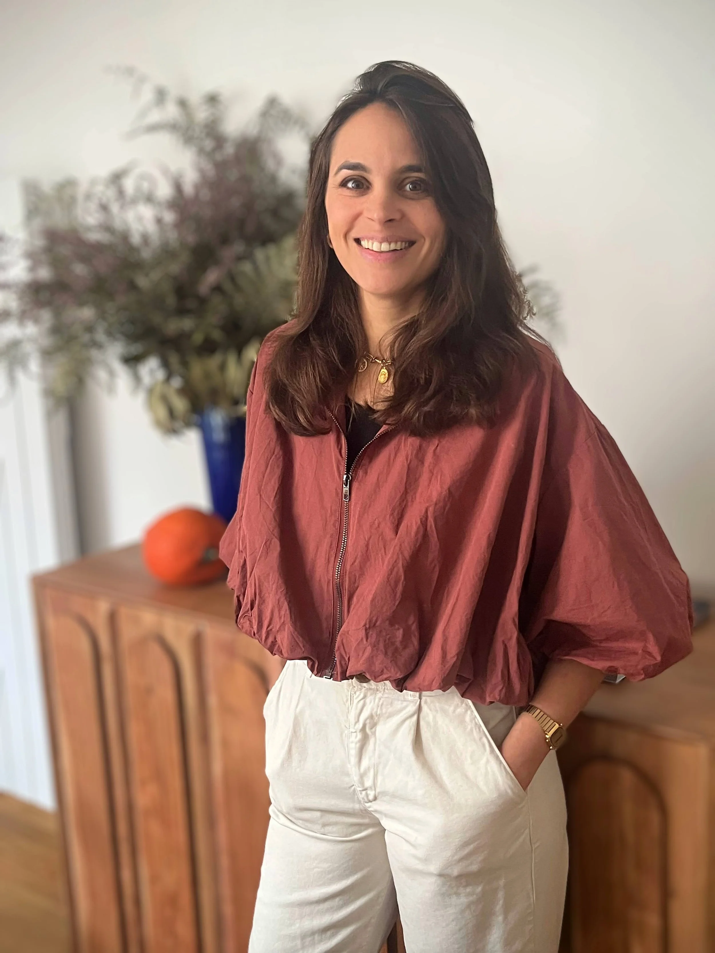 Une femme souriante portant un manteau rouge et un pantalon clair, debout dans une pièce avec un meuble en bois et un bouquet de fleurs en arrière-plan.