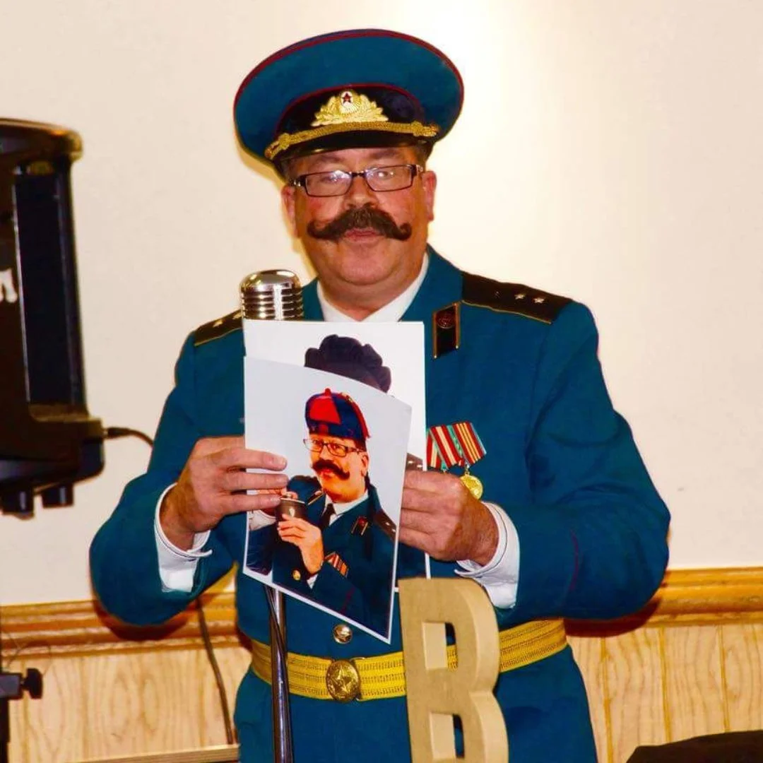 A man dressed in a military uniform with medals and a large mustache, holding a photograph of himself in military attire, standing next to a vintage microphone.