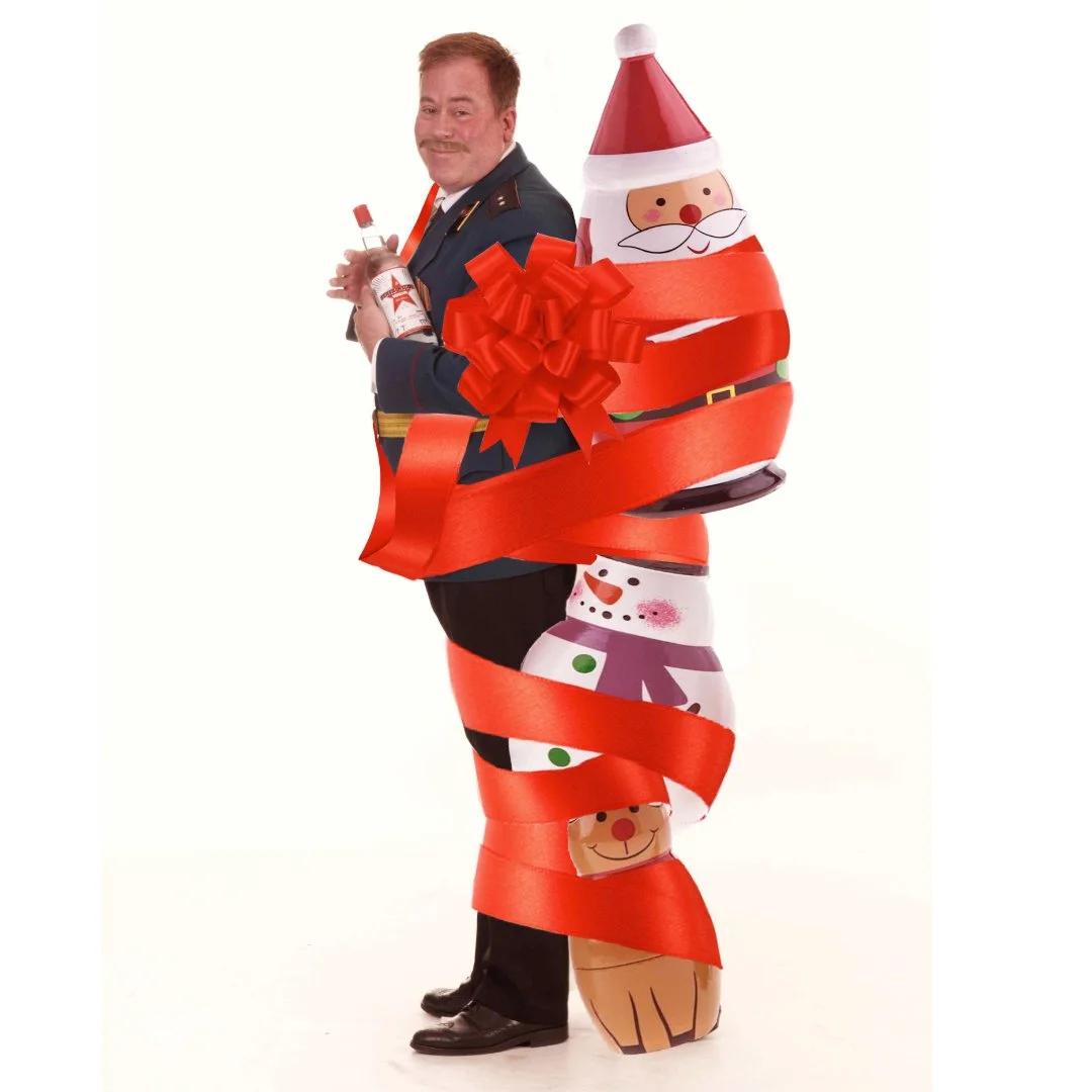 A man dressed in a decorated Santa Christmas costume with large festive ribbons, holding a bottle, and posing with a cheerful smile.