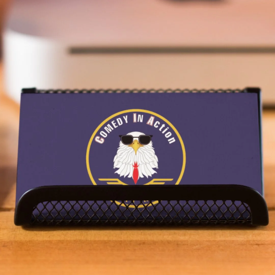Box with a logo of a bald eagle wearing sunglasses, with the text 'Comedy in Action' surrounding it, placed on a black mesh desk organizer on a wooden surface.