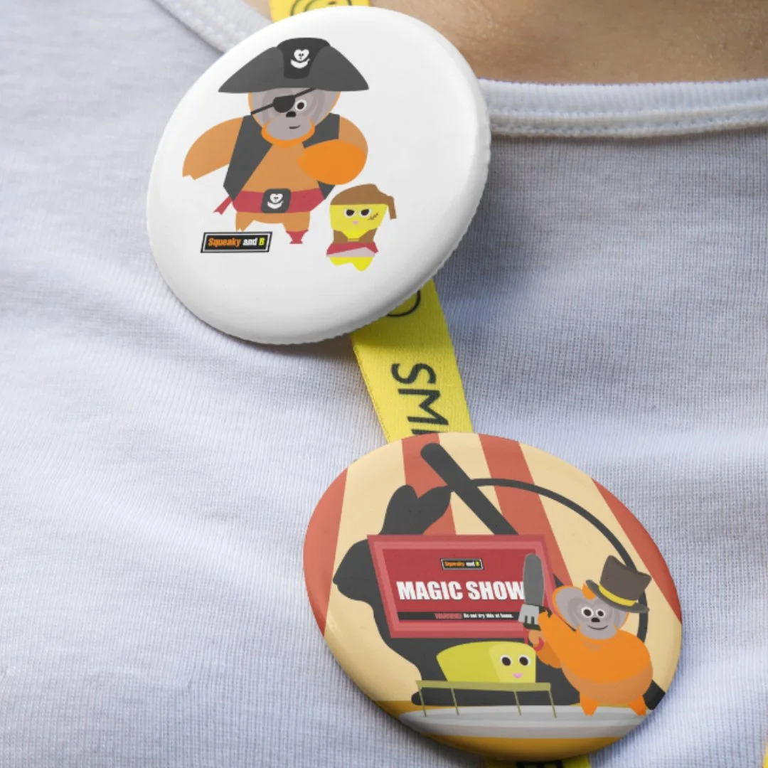 Two buttons with pirate-themed illustrations and text worn on a person's shirt. The top button features a bear dressed as a pirate, holding an orange object, with a smaller yellow character next to it, and the text "Squeaky and B". The bottom button shows a bear with a hat and a sword next to a yellow character, with a sign reading "MAGIC SHOW."