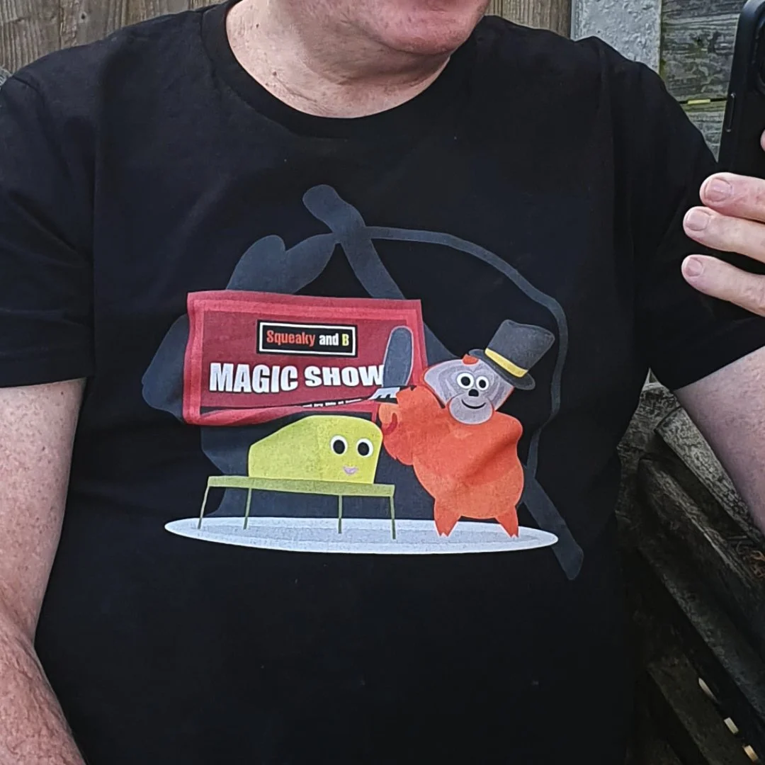 A person wearing a black T-shirt with a cartoon graphic of a magic show featuring a yellow character and gray dog with a top hat, with the words "Squealy and B Magic Show" on a red sign.