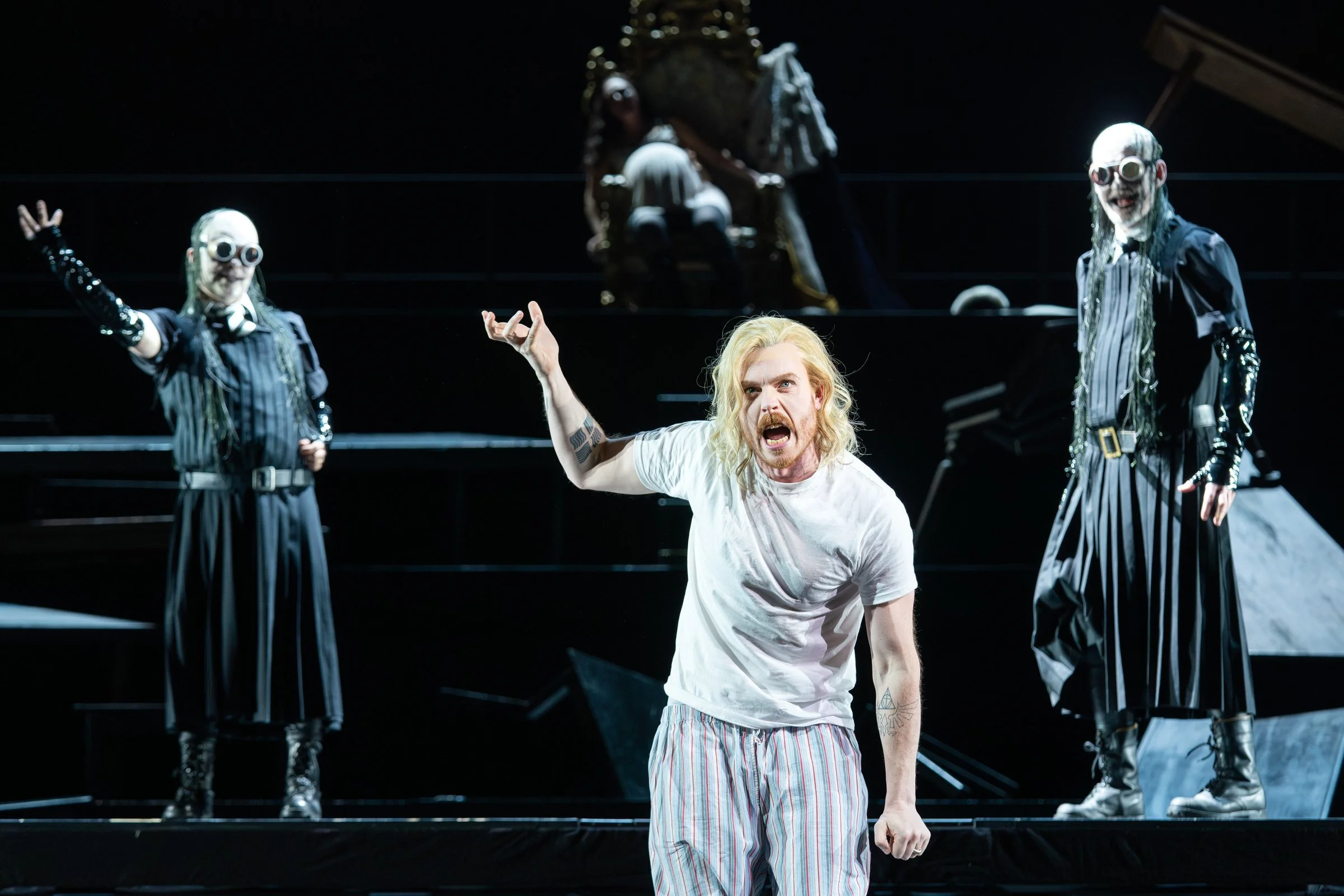 Schauspieler mit langen blonden Haaren und weißen Shirt in einer Theaterszene mit zwei Gestalten in schwarzen Kostümen und weißen Masken im Hintergrund.