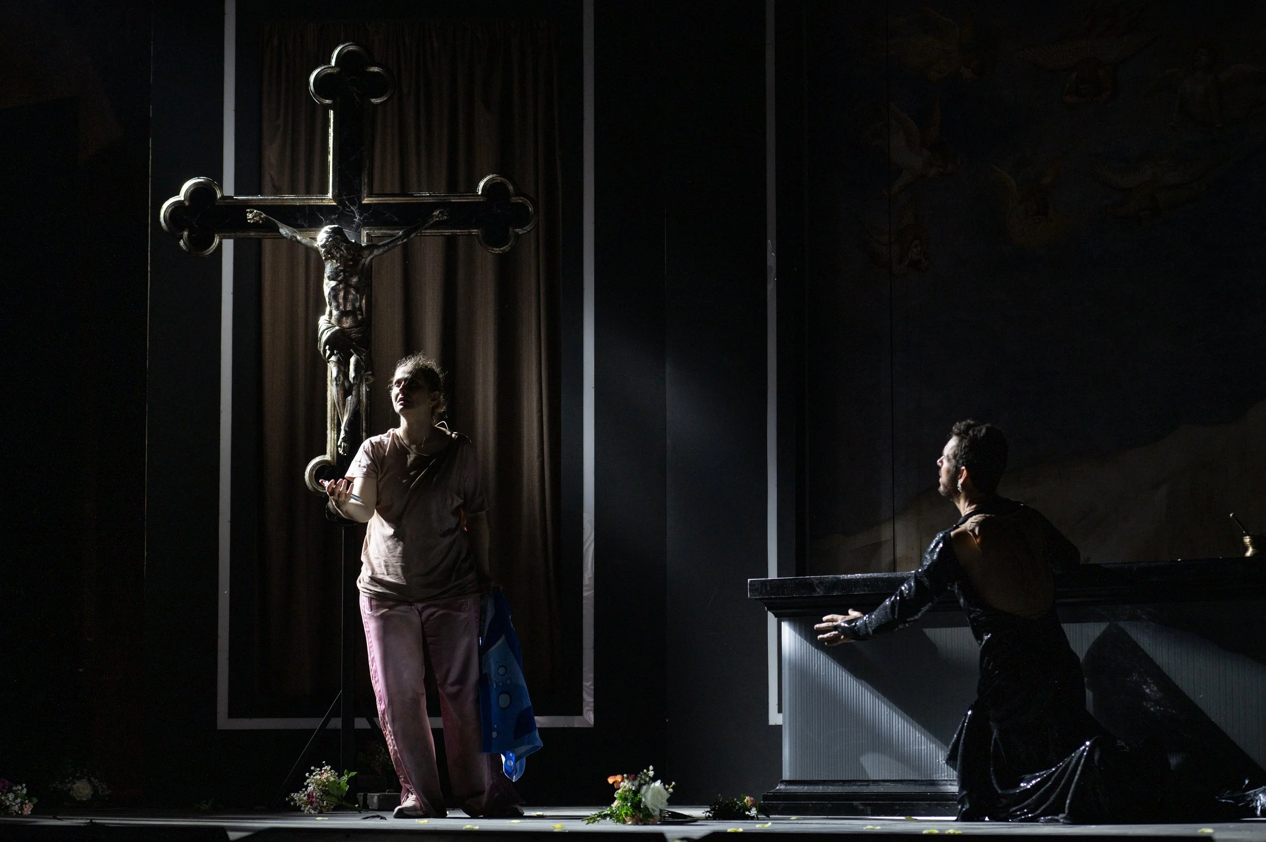 Zwei Personen in der Kirche vor einem großen Kreuz mit Jesus Christus. Eine Person steht und spricht, die andere sitzt auf einer Bank. Es gibt Blumen auf dem Boden.