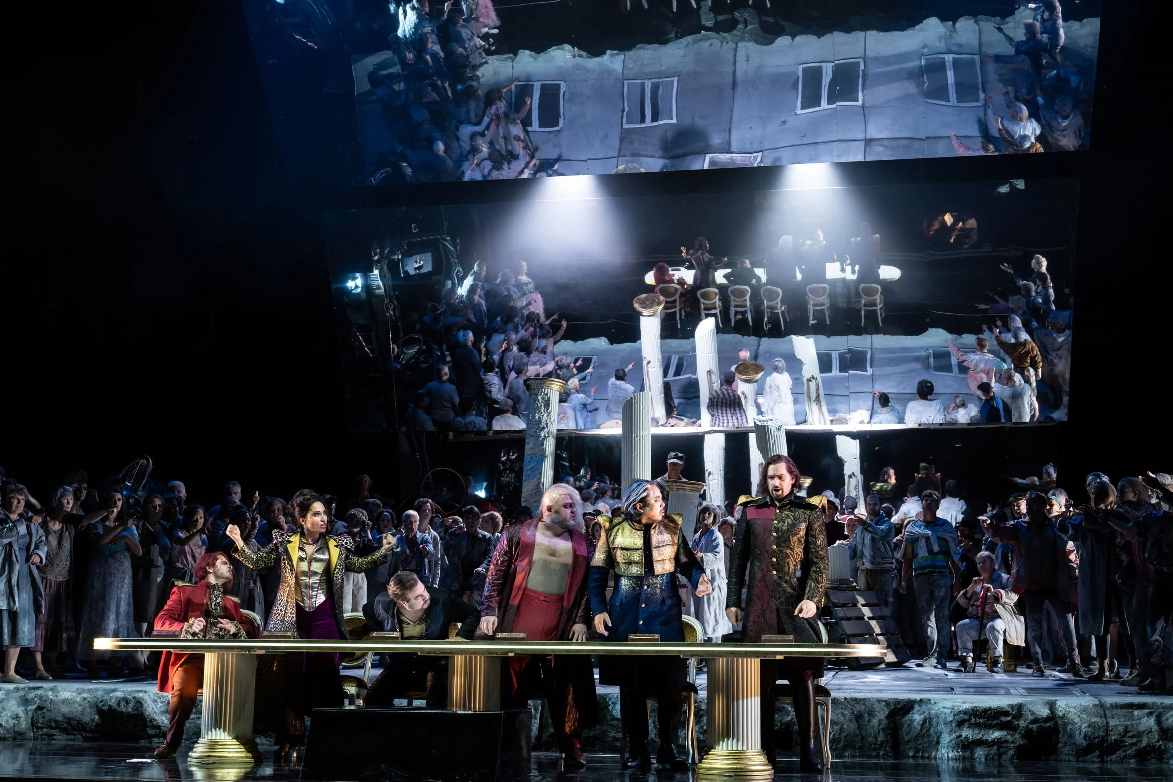 Bühnenszene eines Opern- oder Theaterstücks mit Schauspielern in prunkvollen Kostümen vor einer großen Menschenmenge. Im Hintergrund Spiegel, die die Szene reflektieren.