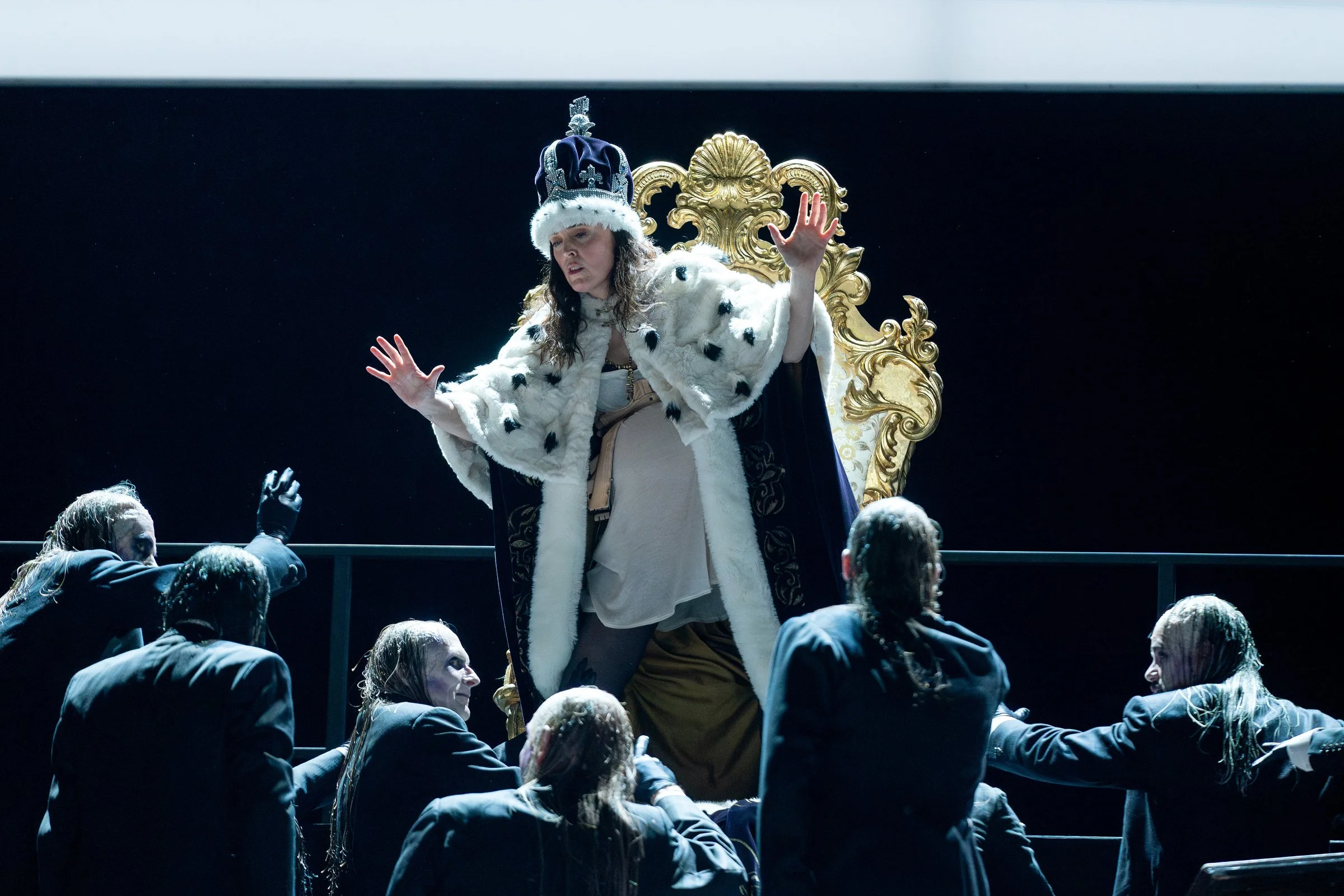 Eine Frau in königlicher Kleidung sitzt auf einem goldenen Thron, umgeben von mehreren Männern in schwarzen Anzügen, die Wasser über ihre Köpfe gießen. Die Szene wirkt dramatisch, mit dunklem Hintergrund und einer Beleuchtung, die die zentrale Figur 