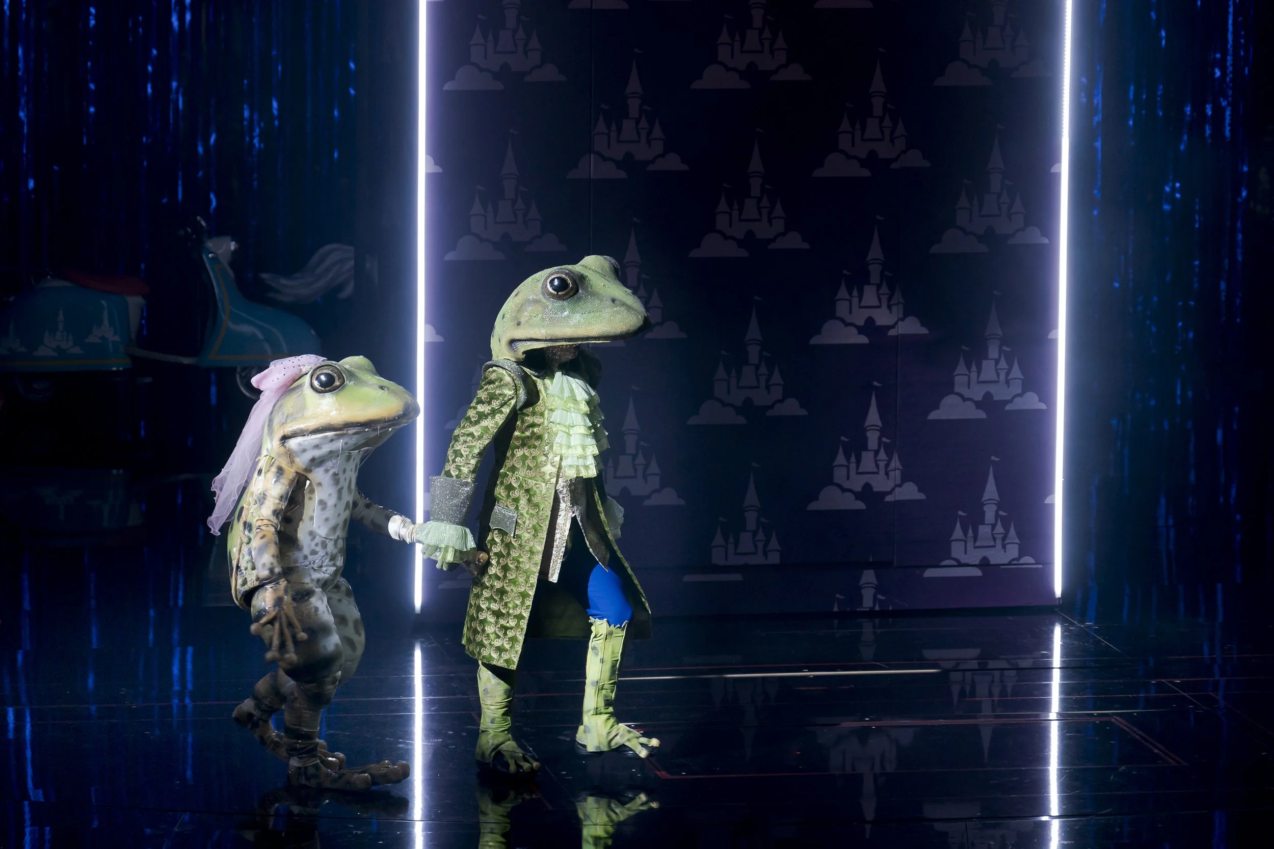 Zwei anthropomorphe Frösche in elegant Kleidung auf einer Bühne mit dunklem Hintergrund und Lichteffekten.