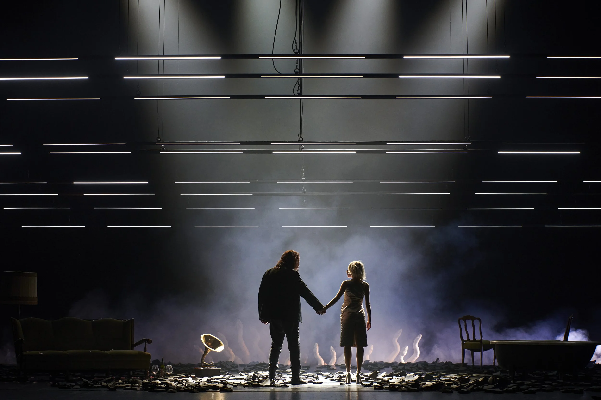 Zwei Personen, ein Mann und eine Frau, halten sich an den Händen und stehen in einem dunklen Raum auf einer Bühne, umgeben von Requisiten und Requisiten, mit dramatischem Licht und Rauch im Hintergrund.