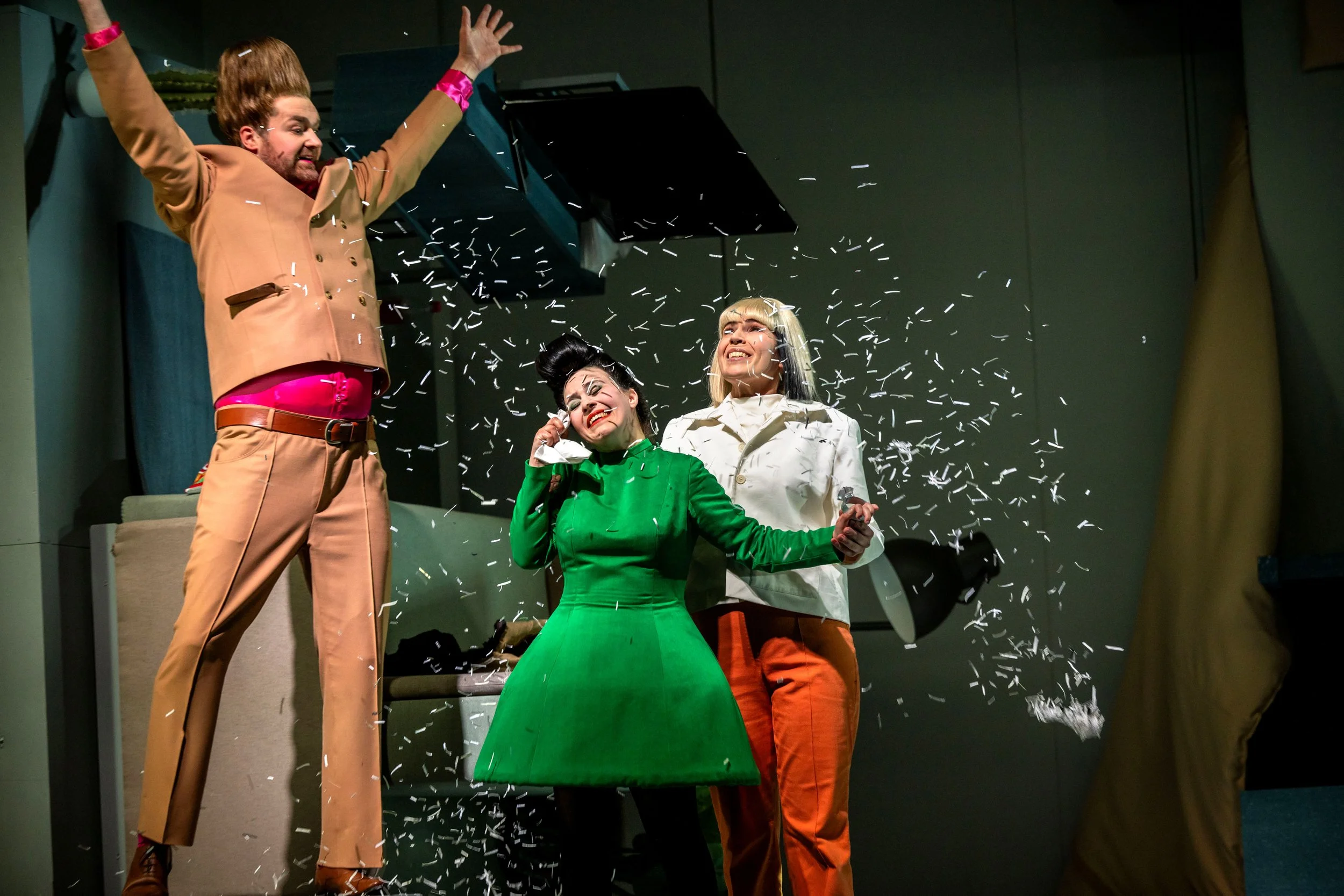 Drei Menschen feiern auf einer Bühne, während Konfetti in die Luft fliegt. Ein Mann wirft mit einem schwarzen Container, während zwei Frauen lachen und sich freuen.
