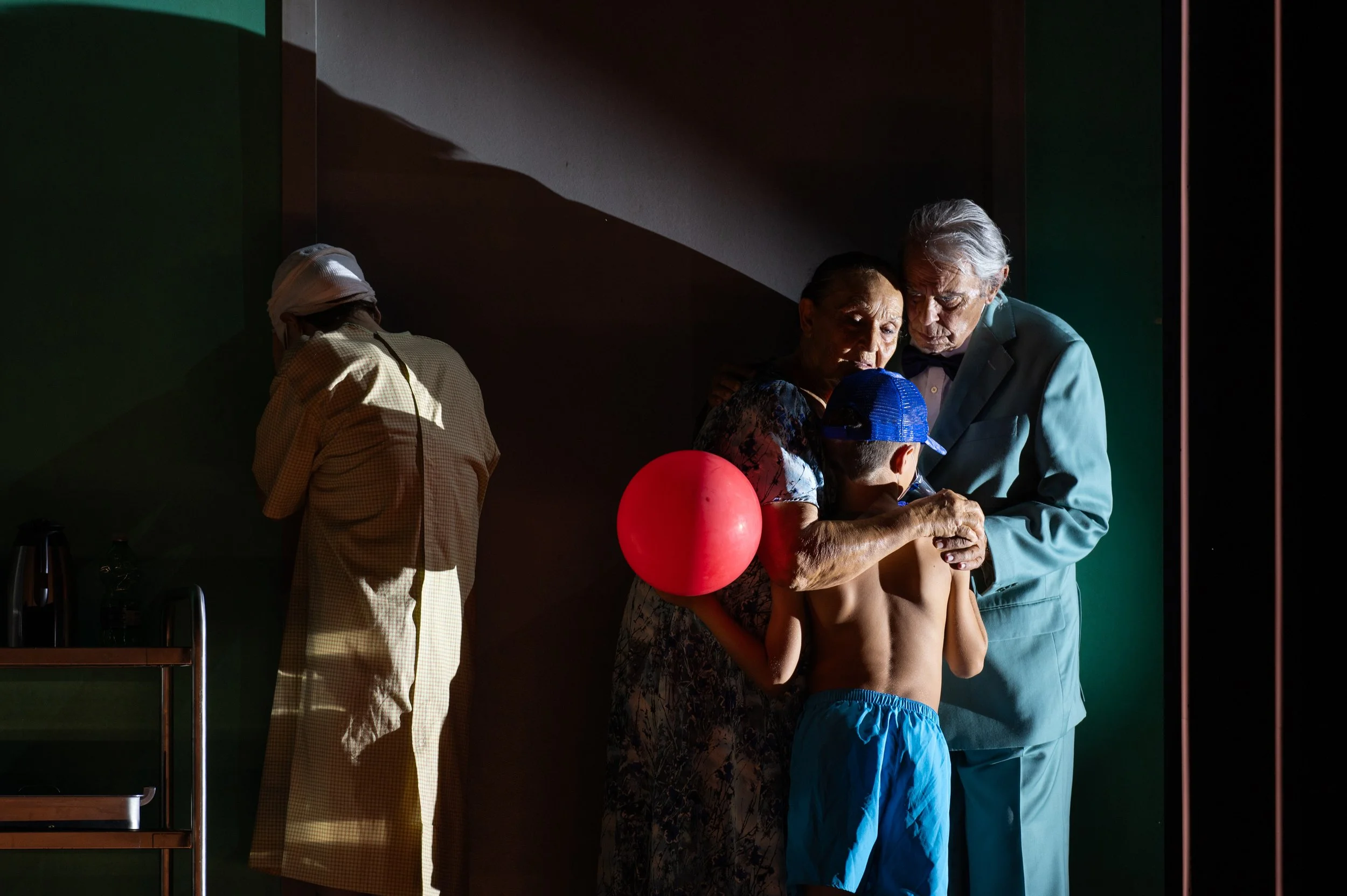 Ein älteres Paar hält einen Jungen mit einem Baseballkappe und einem roten Ball, die beiden scheinen emotionale Momente zu teilen, während eine Frau im Hintergrund sich abwendet.