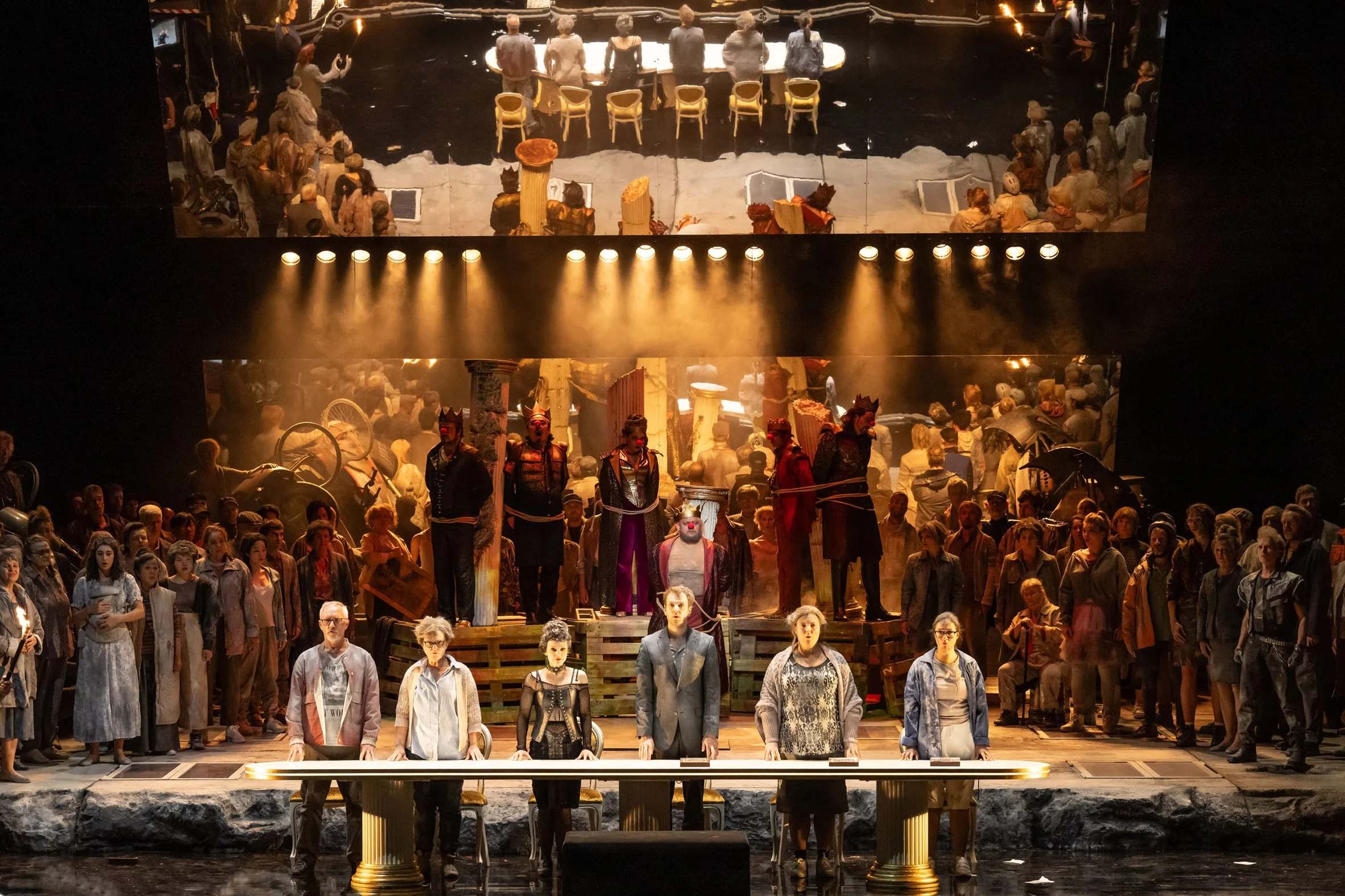 Bühnenaufnahme eines Theaters mit einer großen Gruppe von Schauspielern, die in einer dramatischen Szene stehen. Im Vordergrund stehen sechs Schauspieler an einem langen Pult, einige tragen moderne Kleidung, andere tragen Kostüme und Masken. Im Hinte