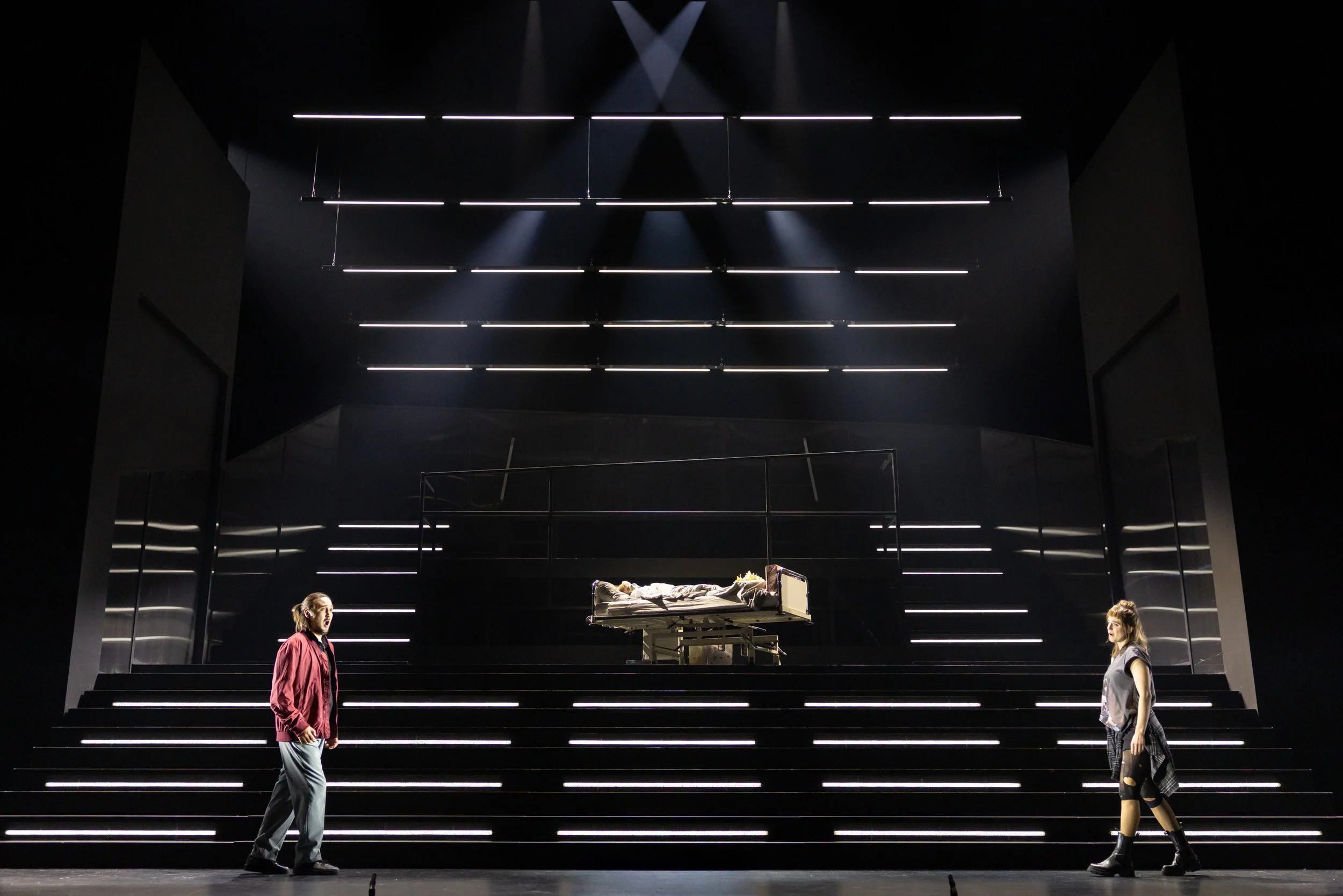 Zwei Schauspieler stehen sich auf einer Bühne mit schwarzen Stufen gegenüber. In der Mitte der Bühne befindet sich ein Krankenbett mit einer liegenden Person. Die Bühne ist dunkel mit horizontalen Lichtstreifen an Wänden und Decke.