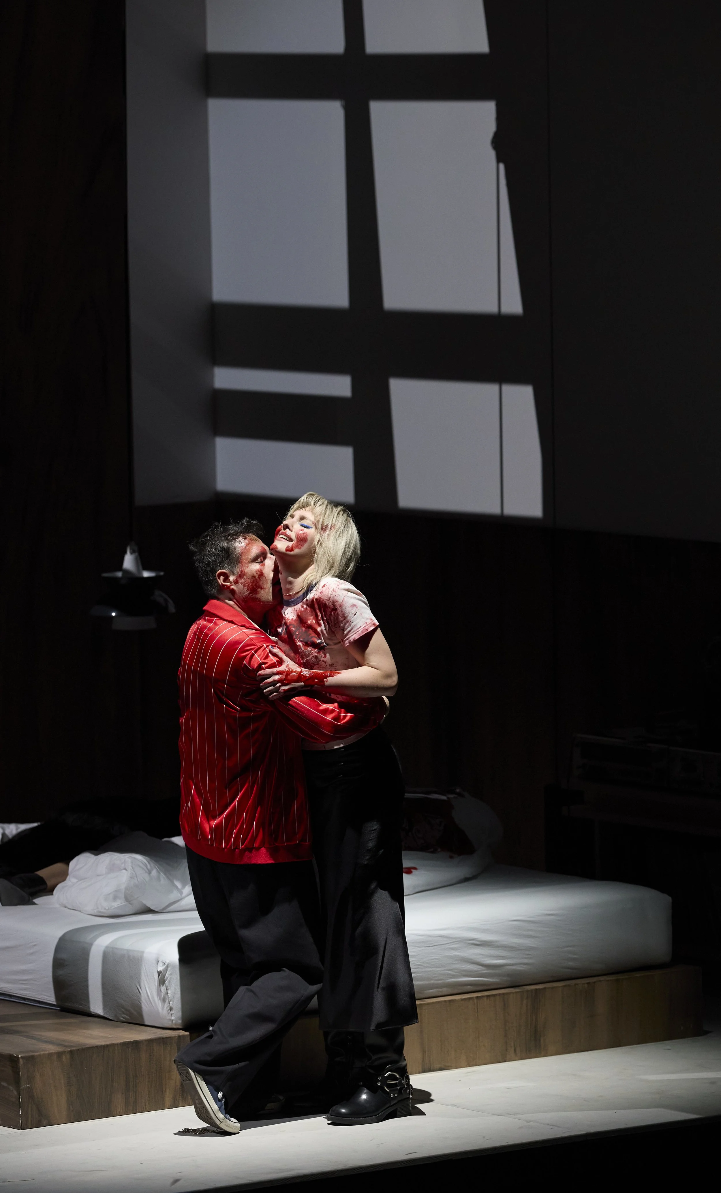 Zwei Menschen umarmen sich auf einer Bühne, eine Person trägt eine blutverschmierte Kleidung, im Hintergrund ein Fenster mit Schatten. Ein Bett ist im Raum sichtbar.