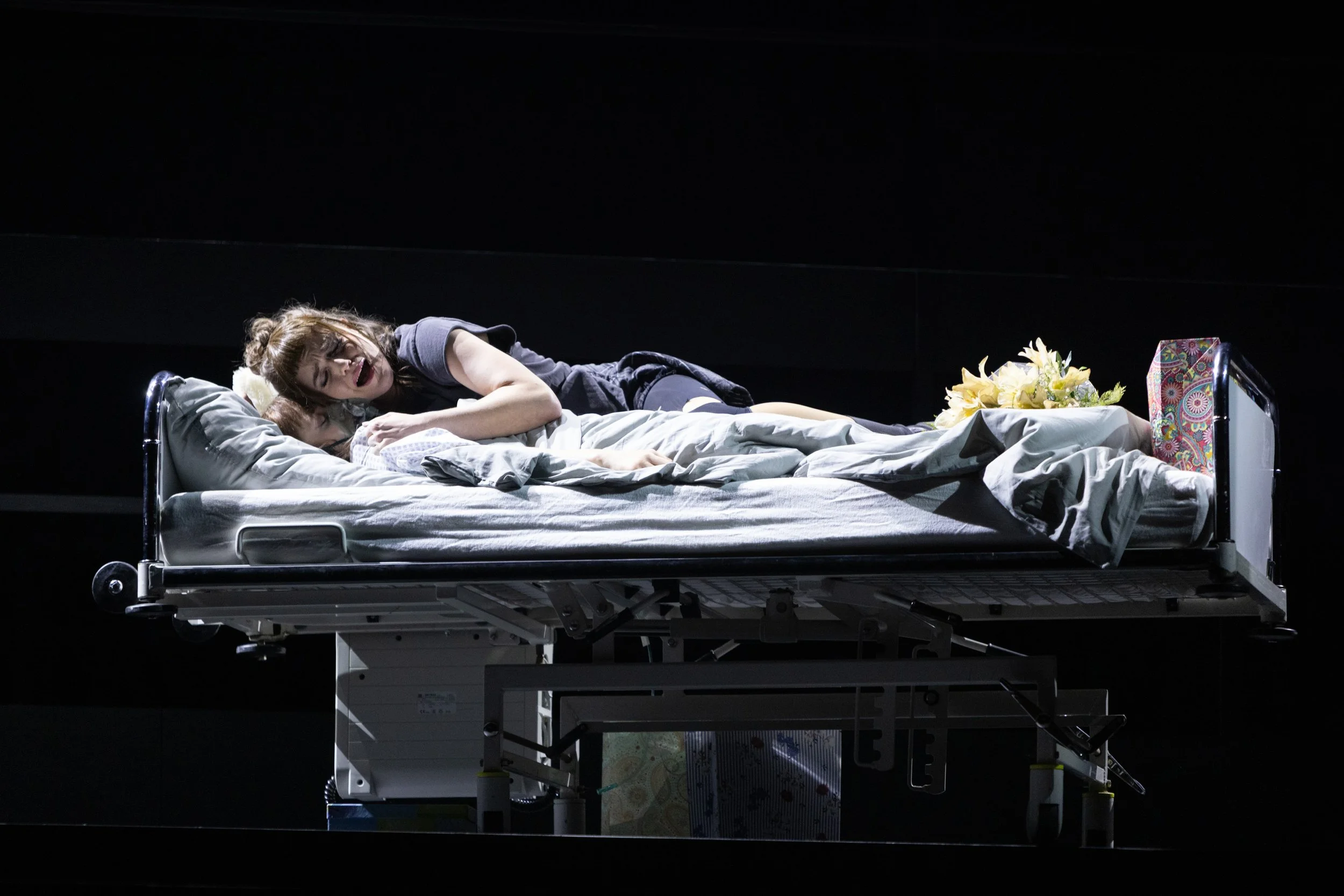 Szene auf einem Krankenhausbett: Eine Frau liegt auf einer Reihe mit weißen Bettwäsche, offensichtlich im Gespräch mit einer anderen Frau. Im Hintergrund befinden sich Blumen und eine Geschenkbox.