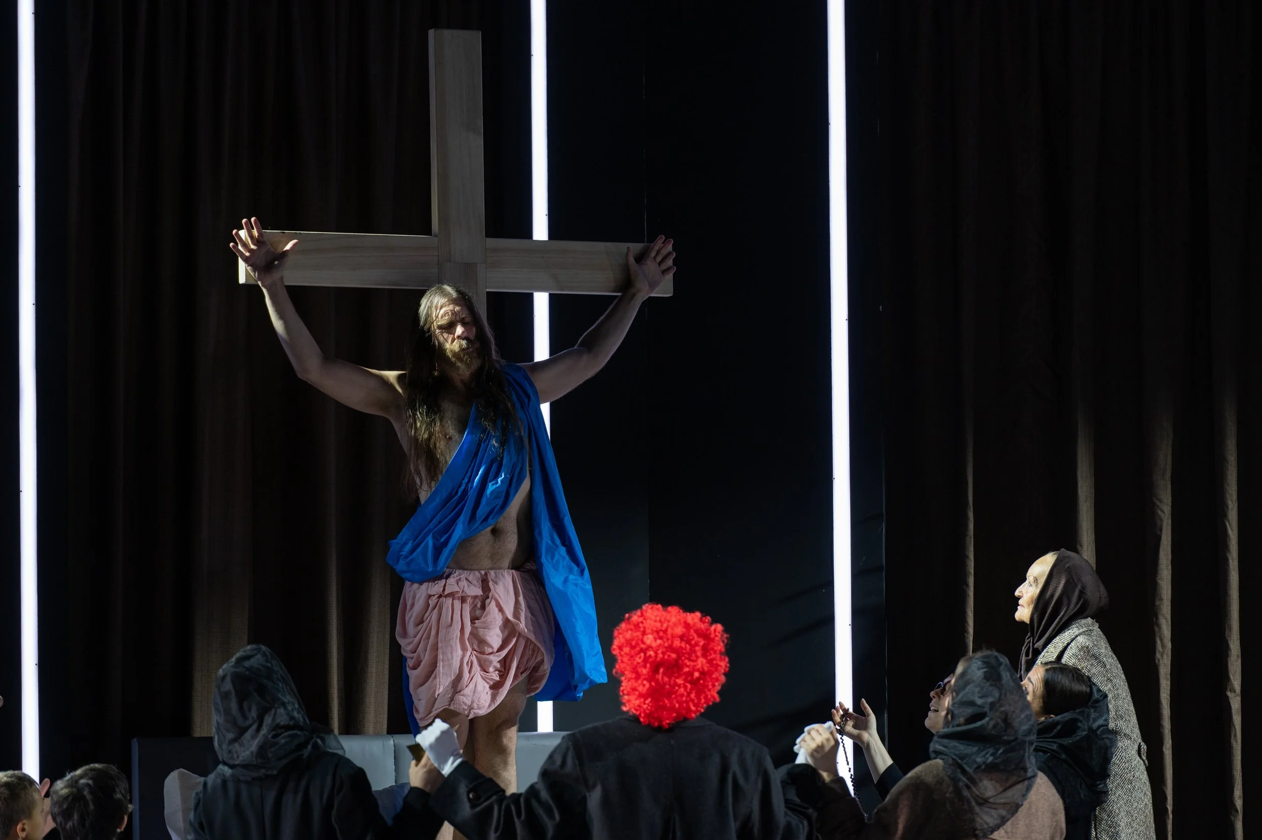 Eine Person, die Jesus Christus als Figur mit einem Holzkreuz auf den Schultern darstellt, steht auf einer Bühne vor einer Gruppe von Zuschauern in dunkler Kleidung. Die Szene wirkt wie eine Theateraufführung.