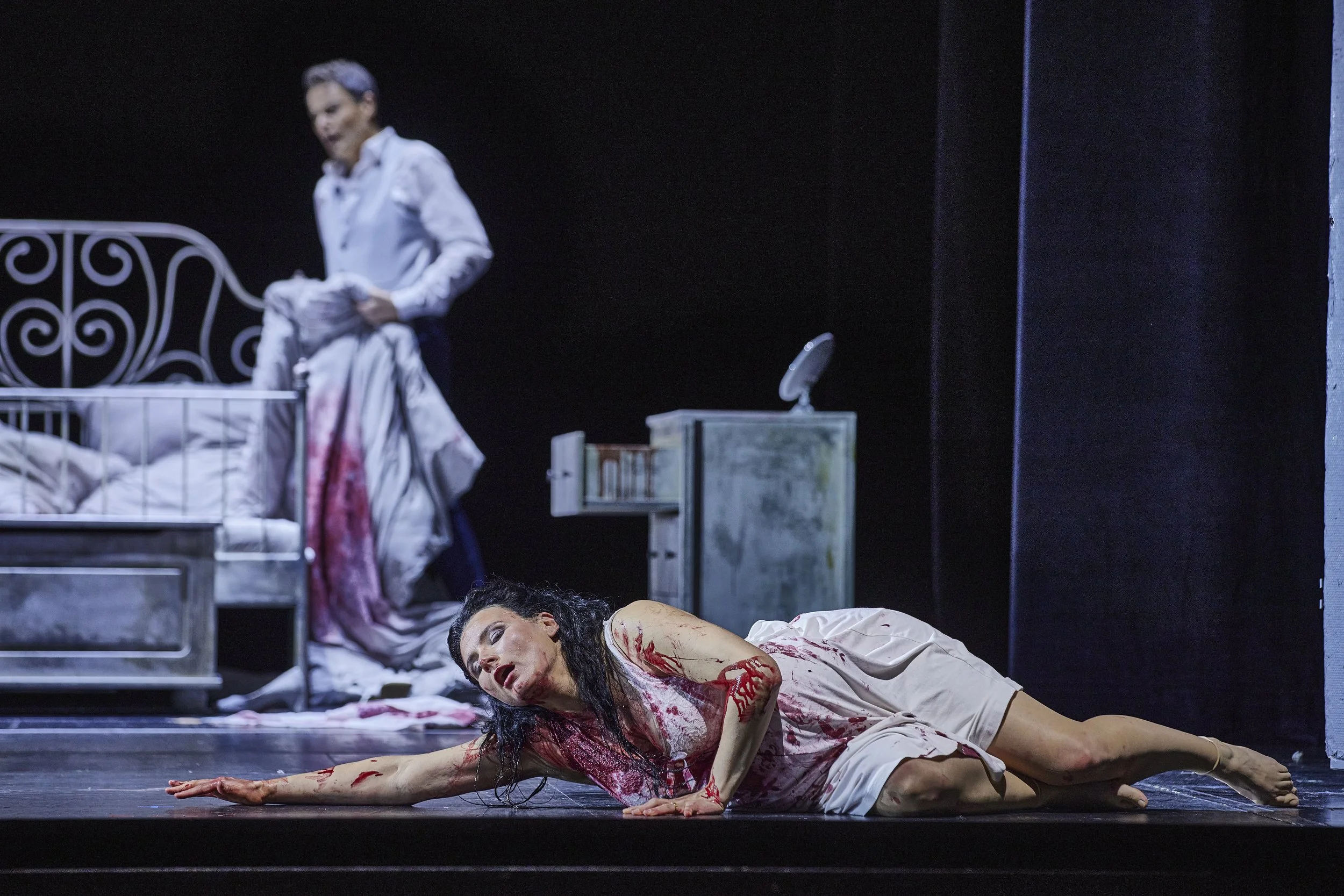 Szene aus einem Theaterstück mit einer verletzten Frau, die auf dem Boden liegt, während eine andere Person im Hintergrund bei einem Bett steht.