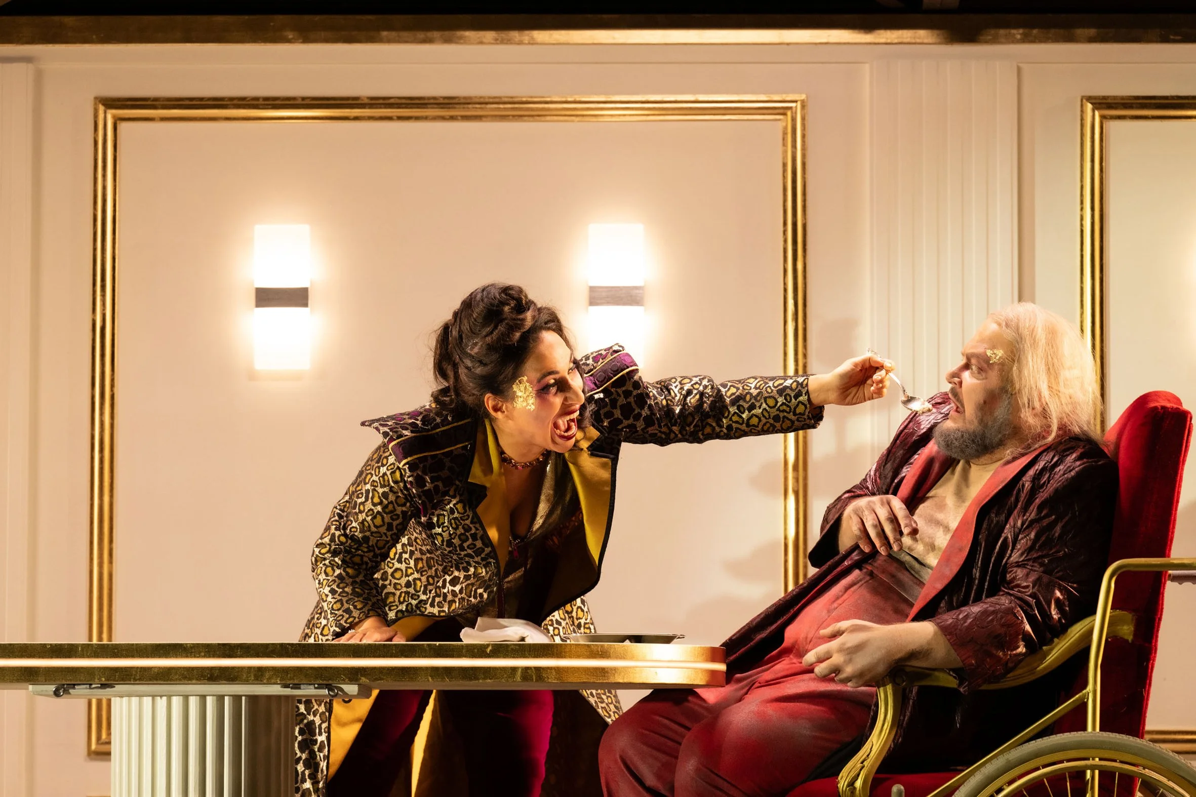 Eine Frau in einem auffälligen, gemusterten Outfit füttert einen älteren Mann im Rollstuhl mit einem Löffel in einem eleganten Raum mit goldenen Akzenten.
