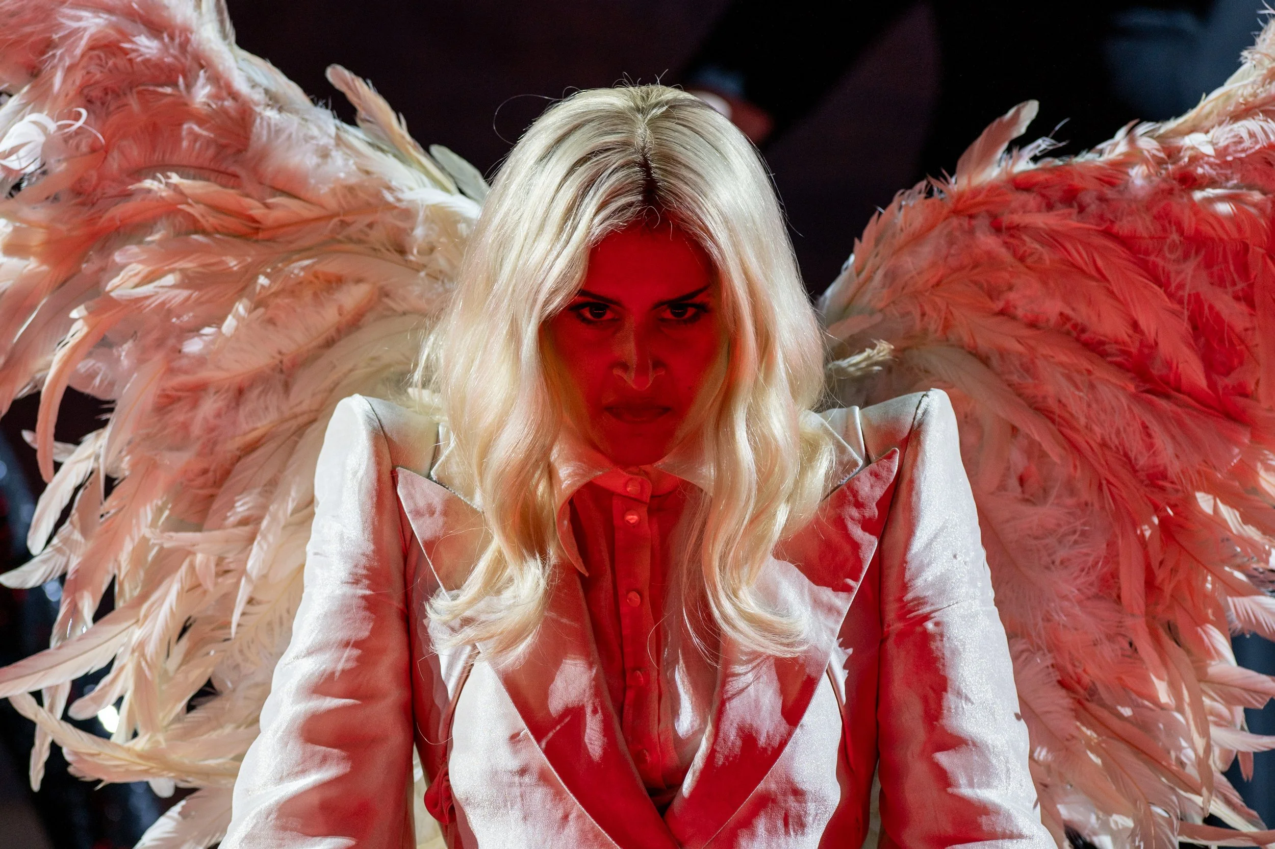 Frau mit großen rosa Flügeln im weißen Anzug und rotem Hemd, emotionale Mimik, dunkler Hintergrund.