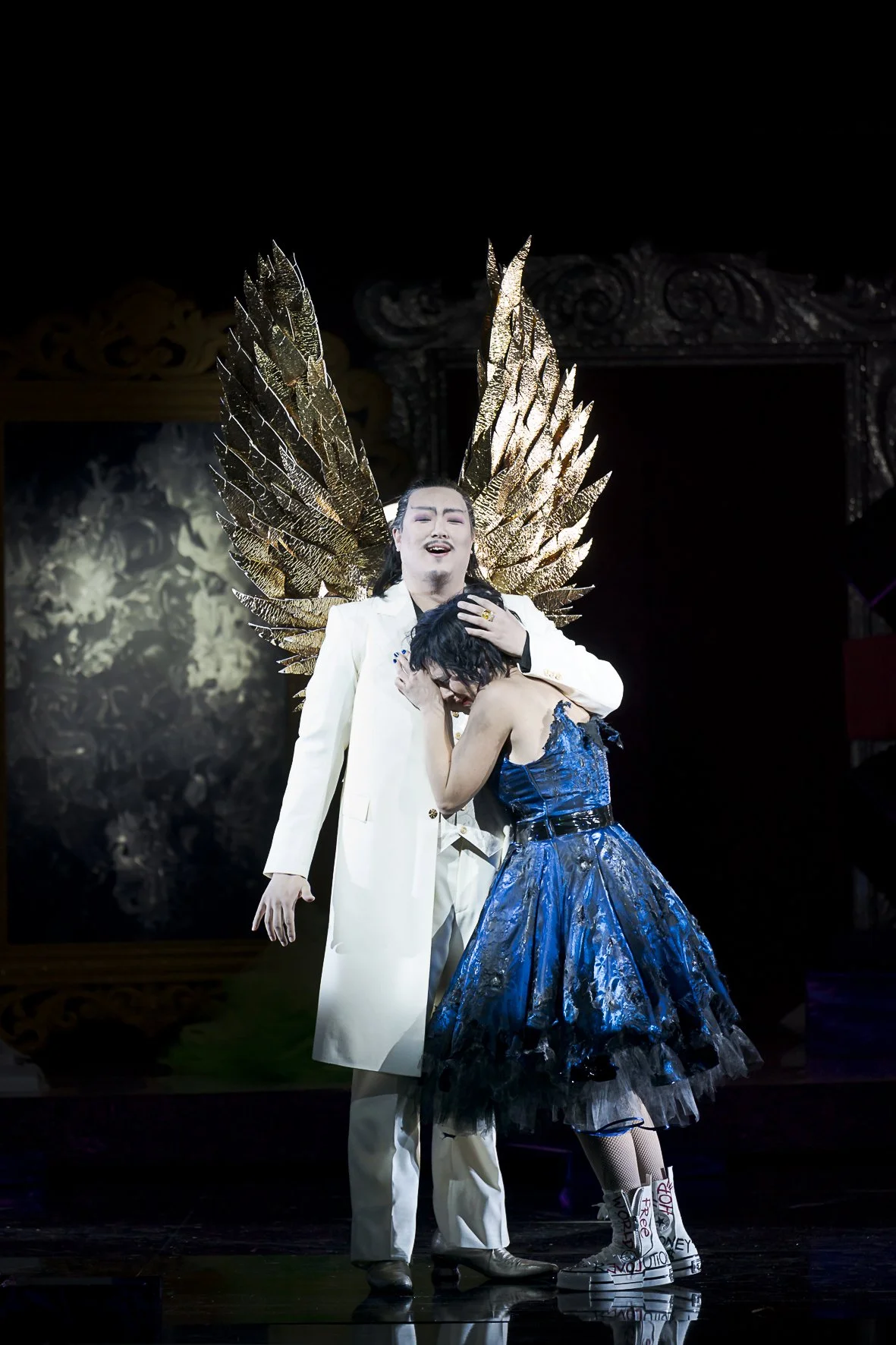 Theateraufnahme mit zwei Schauspielern, einer trägt große goldene Flügel, der andere ist umarmt und weint.