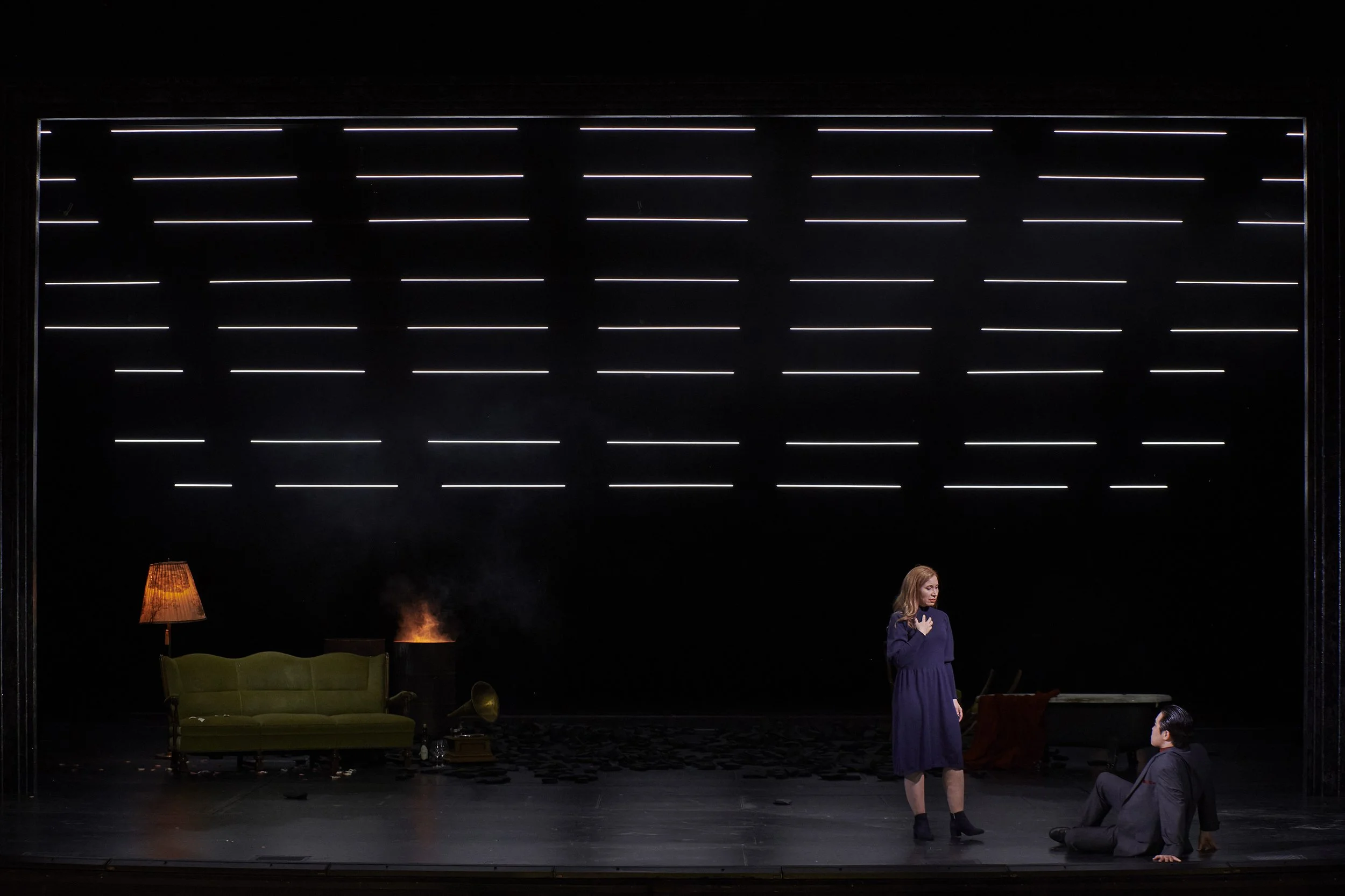 Zwei Schauspieler auf einer Bühne, eine Frau steht und ein Mann sitzt am Boden, im Hintergrund ein Sofa mit einer Stehlampe und einem schimmernden Gegenstand, beleuchtet durch eine Kulisse mit horizontalen Neonschlägen.