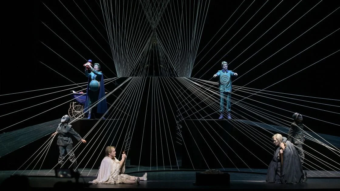 Bühnenaufnahme eines Theaterstücks mit Schauspielern in Kostümen, umgeben von Linien, die wie eine skurrile Szene aussehen.