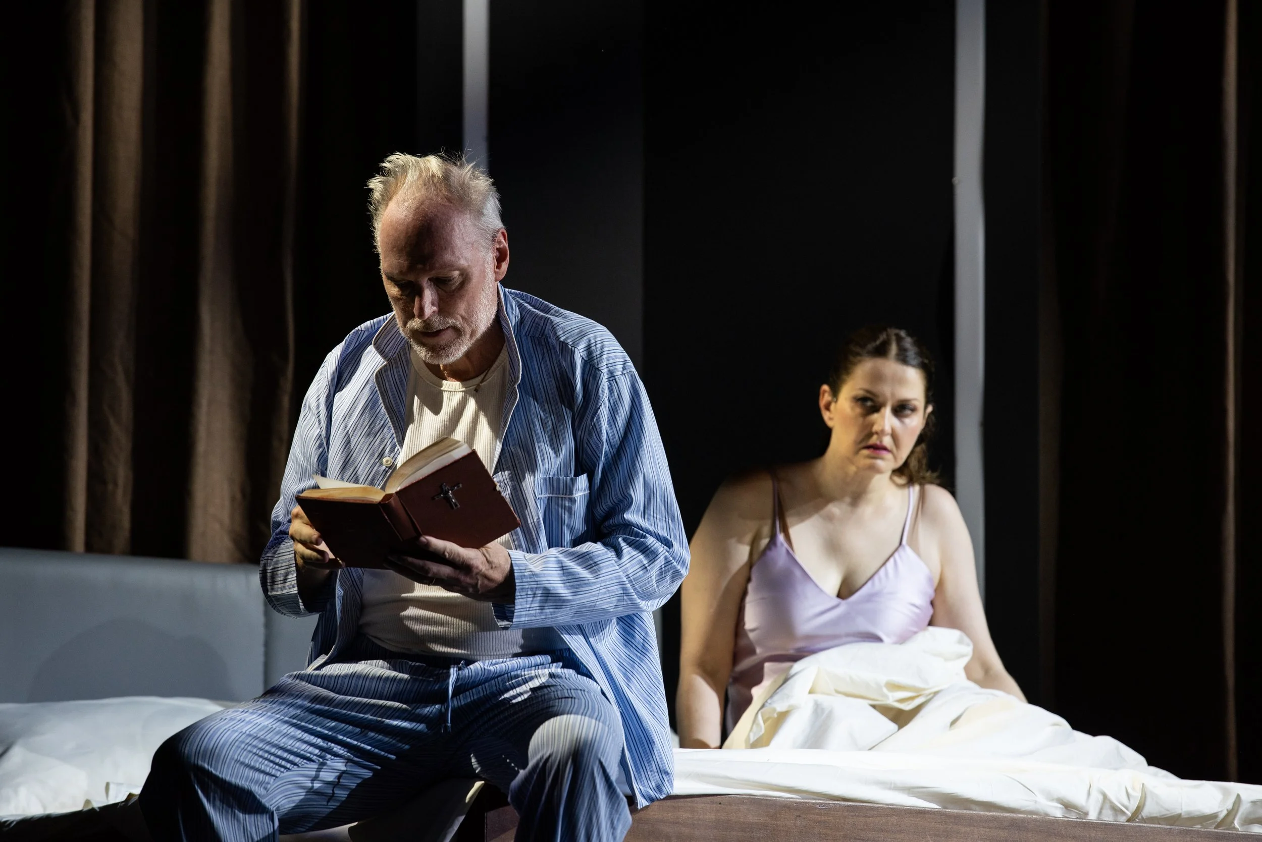 Ein Mann sitzt auf der Bettkante und liest in einer Bibel, während eine Frau im Bett sitzt und ihn beobachtet.