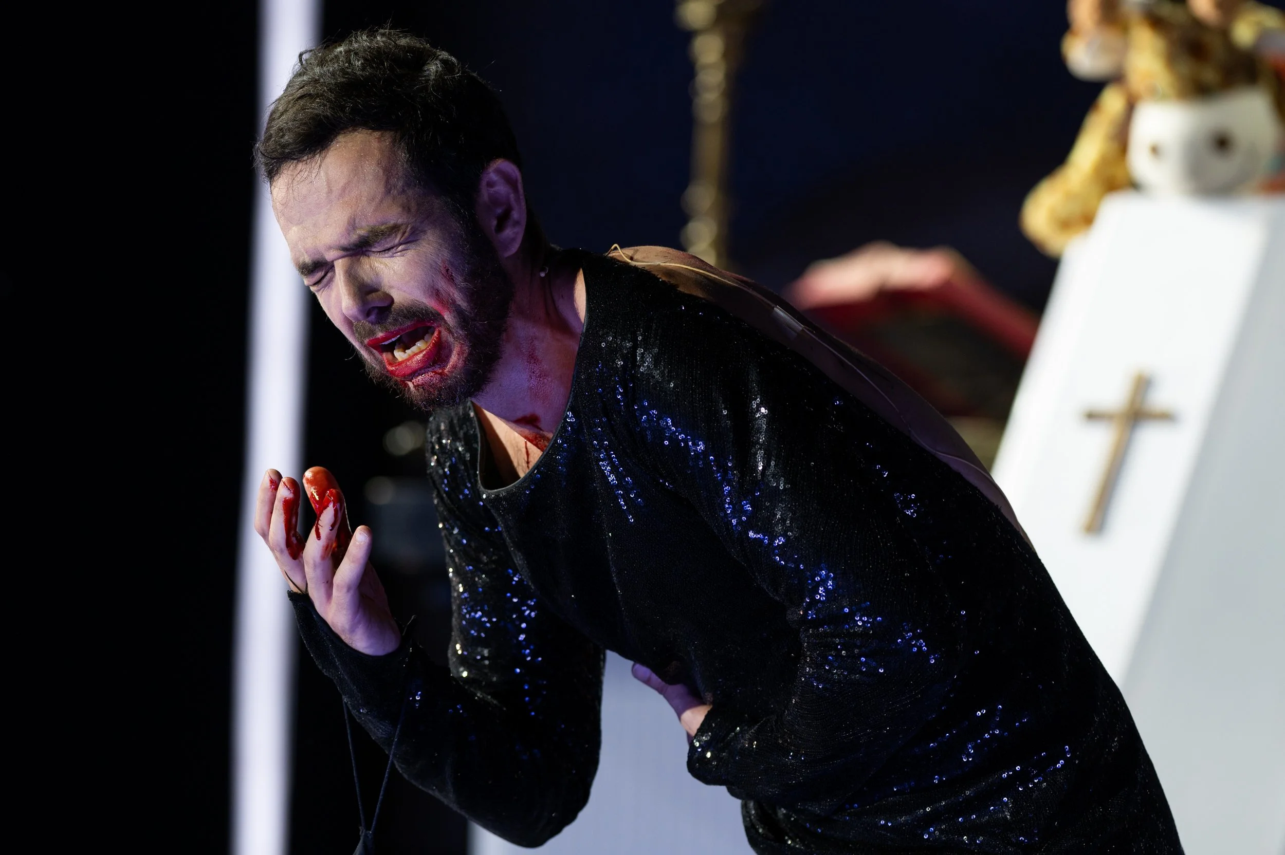 Ein Mann mit blutigem Gesicht und blutigen Händen, der vor Schmerz schreit, in einem schwarzen, glitzernden Outfit, im Hintergrund ein Kreuz und religiöse Gegenstände.