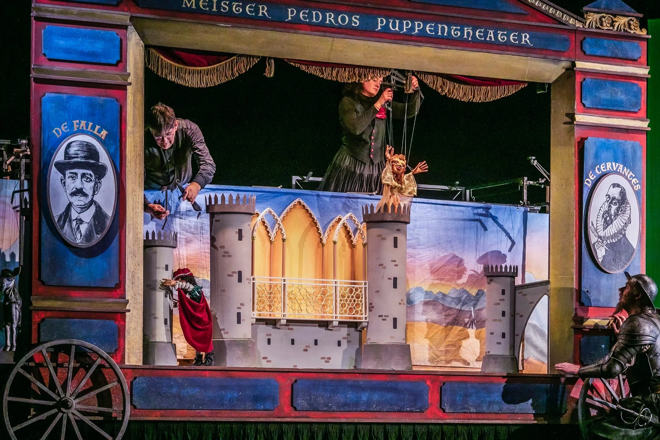 Puppentheater mit mittelalterlichem Schlossmotiv, zwei Bühnenfiguren, eine auf der Bühne und eine im oberen Teil, und Darstellern, die das Stück steuern. Die Kulisse zeigt Berge und Himmel im Hintergrund.