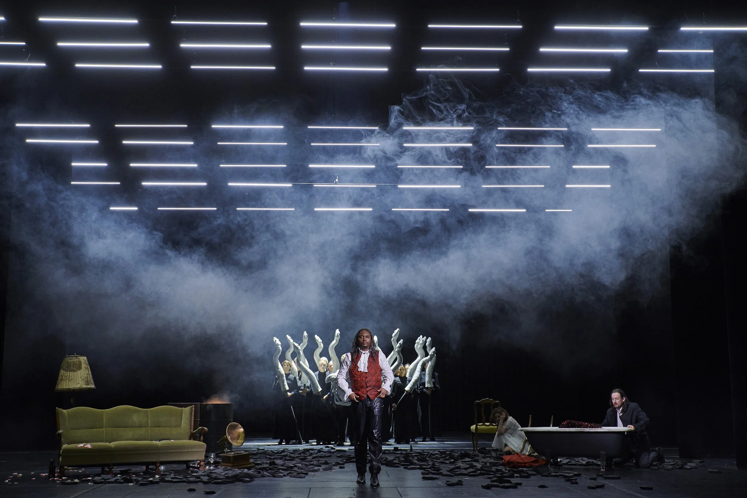 Bühnenszene mit einer Person in der Mitte, umgeben von anderen im Hintergrund, Requisiten und Szenelementen wie Sofa, Lampe und Badewanne, mit Rauch und Neonlichtlinien im Hintergrund.