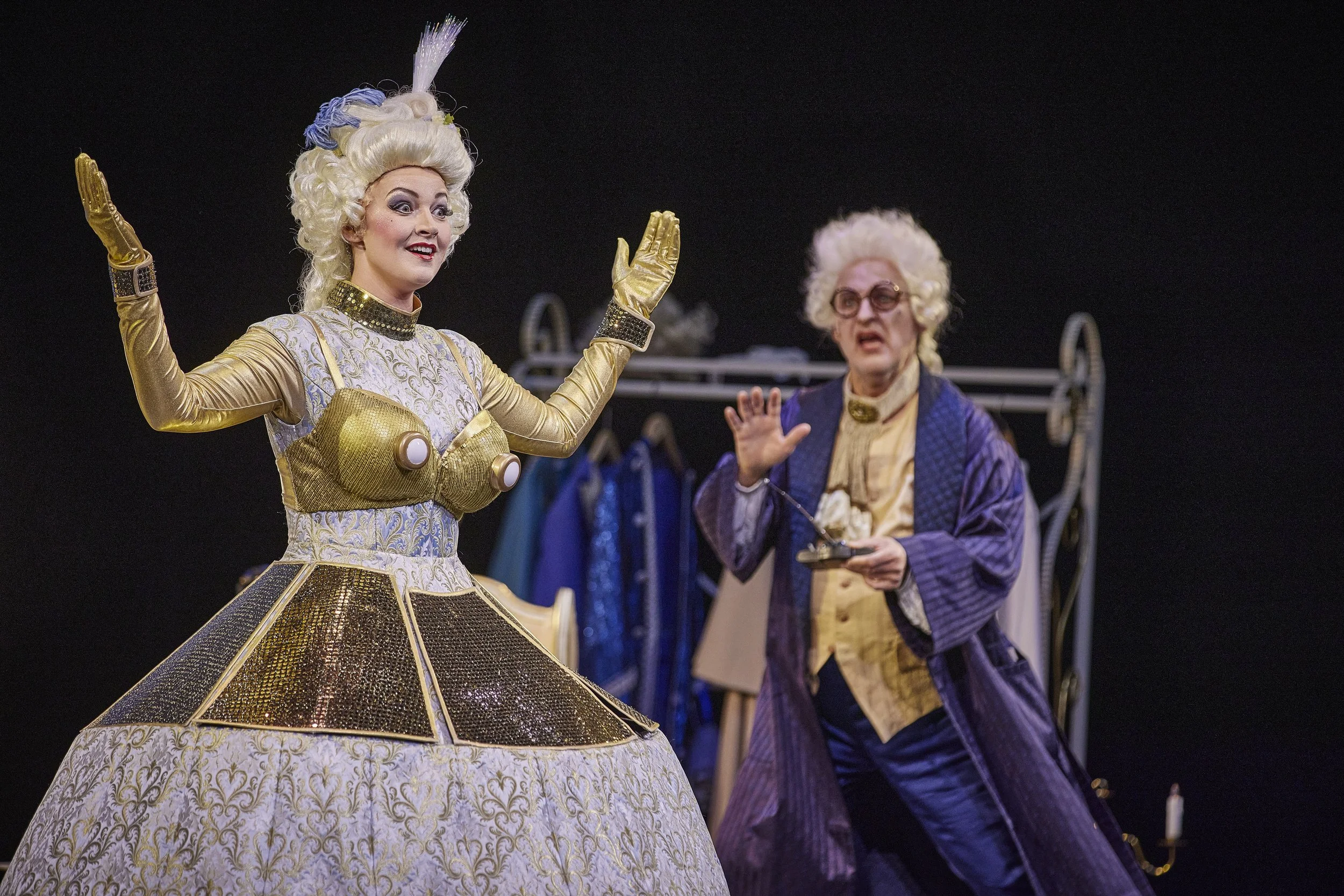 Zwei Schauspieler in aufwändigen, historischen Kostümen, eine Frau in einem gold-glänzenden Kleid und eine ältere Mann in einem Samt-Kostüm, spielen eine Szene auf einer Theaterbühne.