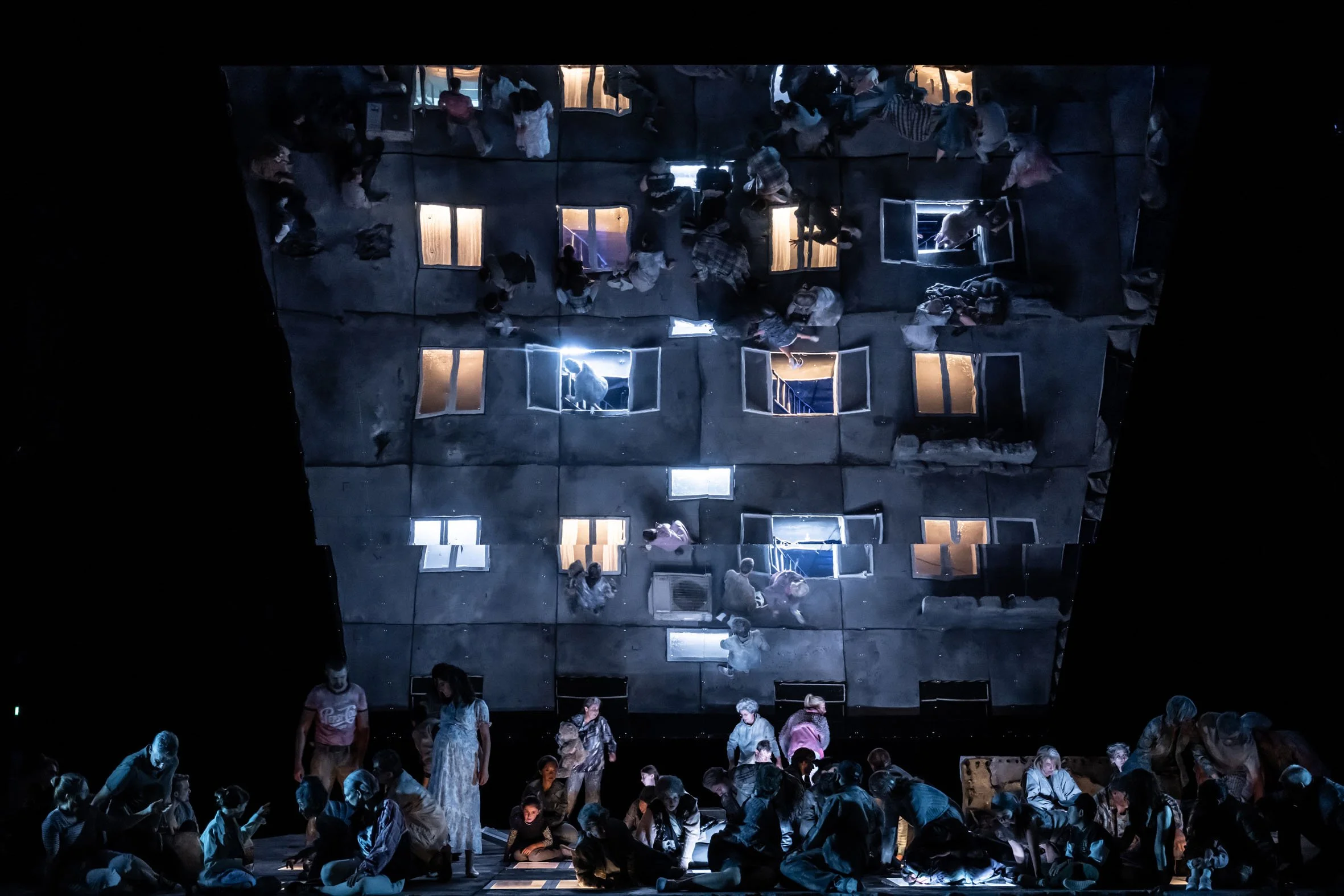 Bühnenbild einer Theateraufführung mit einem großen, vierstöckigen Gebäude, das im Hintergrund schwebt. Das Gebäude hat offene Fenster, durch die Licht und Szenen sichtbar sind. Auf der Bühne sitzen und stehen viele Schauspieler in einer dunklen Umgebung, einige in Gruppierungen, andere allein, mit Beleuchtung auf einzelne Darsteller und das Gebäude im Hintergrund.