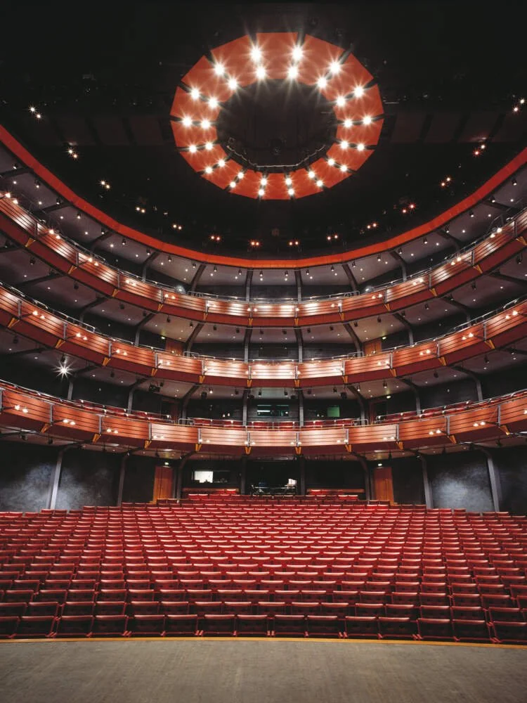 Leerer Theaterraum mit roten Sitzen und mehreren Balkonen, Blick nach vorne zur Bühne, Deckenbeleuchtung und runde Decke