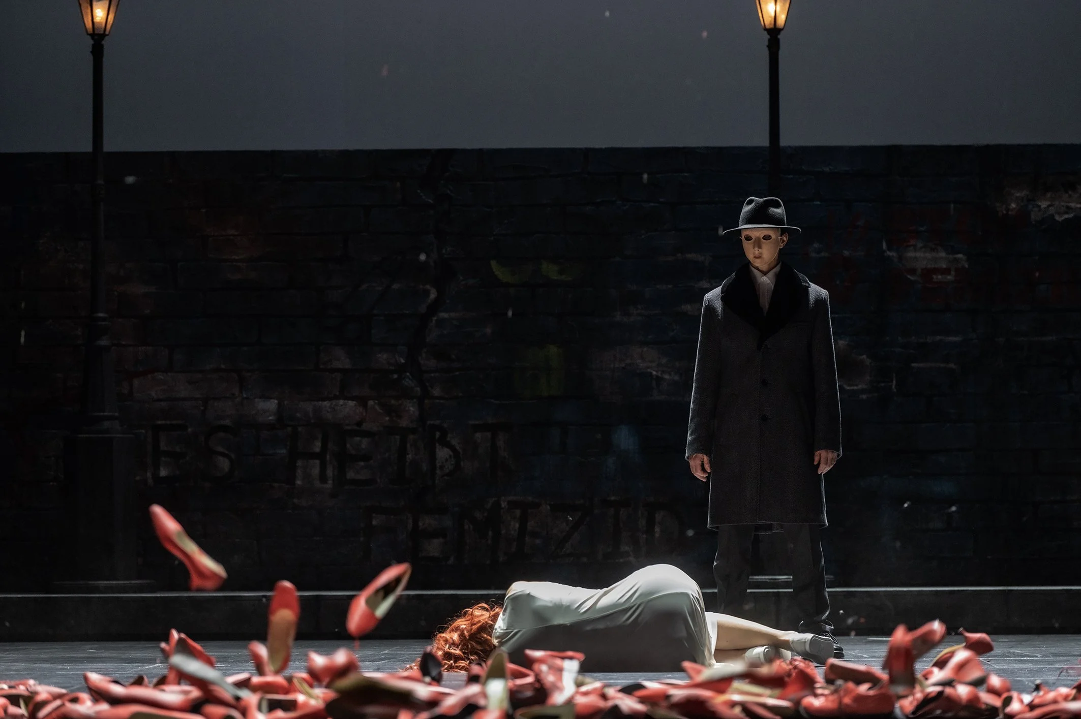 Theaterbühne mit zwei Darstellern, einer liegt auf dem Boden, der andere steht. Der auf dem Boden liegende trägt ein weißes Kleid und rote Schuhe, die im Flug sind. Der stehende trägt einen dunkelgrauen Mantel und Hut, maskiert mit einem weißen Masken, und beobachtet die Szene.