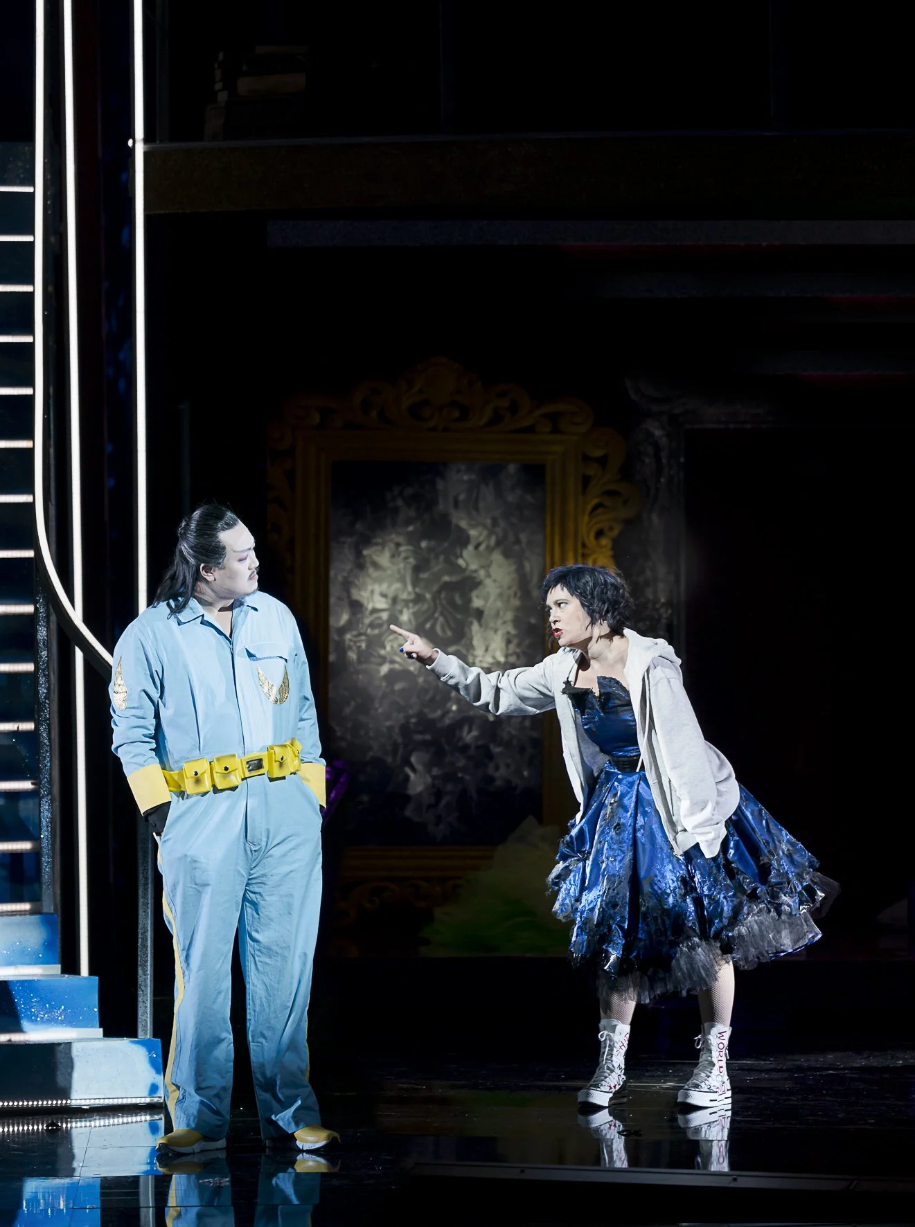 Zwei Schauspieler auf einer Theaterbühne. Der Mann trägt einen blauen Anzug mit Gelbbändern, die Frau trägt ein blauschwarzes Kleid, eine graue Jacke und weiße Turnschuhe mit Text. Die Frau scheint zu dominieren, während der Mann zuhört.