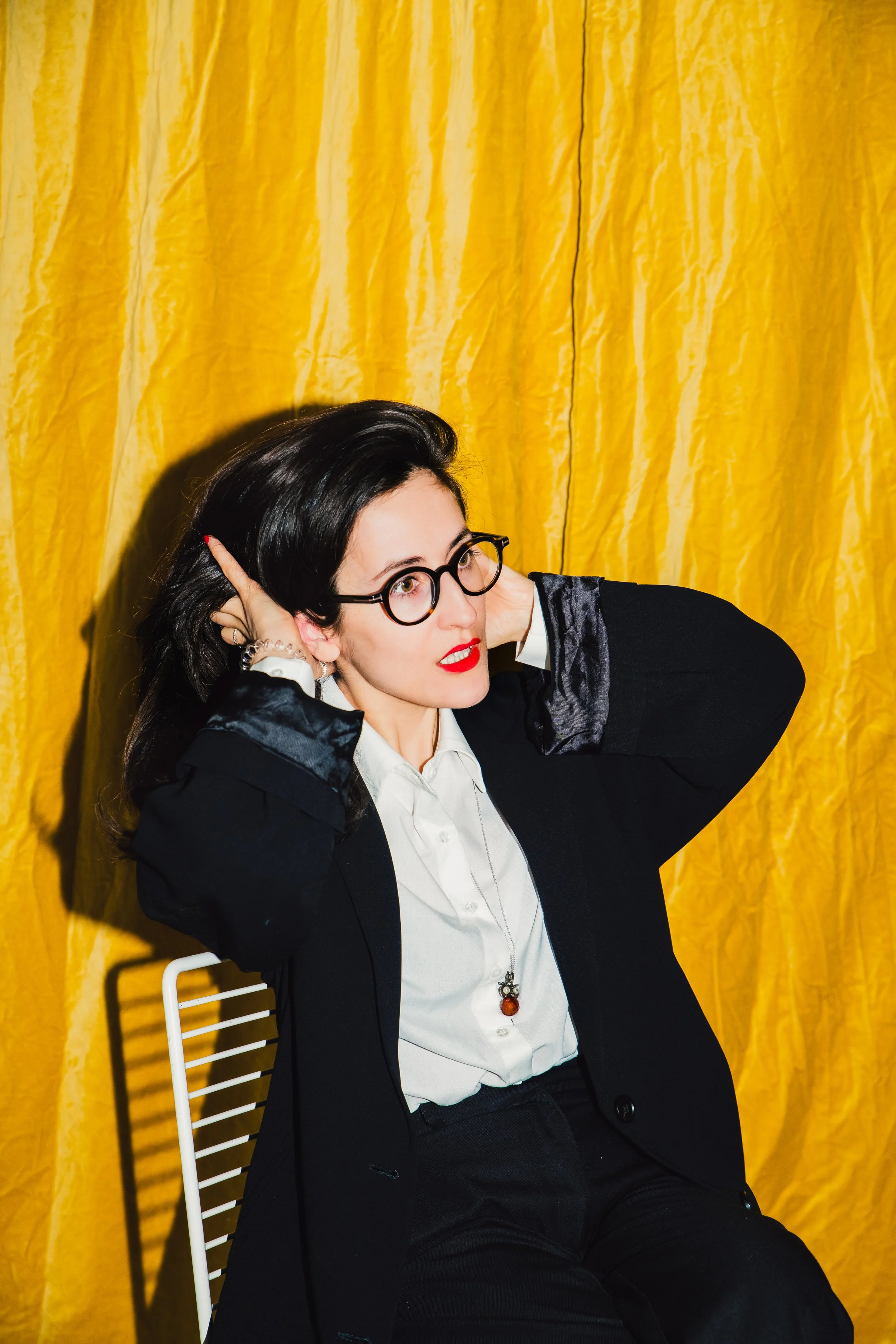 Frau mit schwarzen Haaren und Brille, trägt schwarzen Blazer, weiße Bluse und rote Lippen, sitzt vor gelbem Hintergrund, schaut nach oben, hat die Hände im Haar, sitzt auf einem weißen Stuhl.