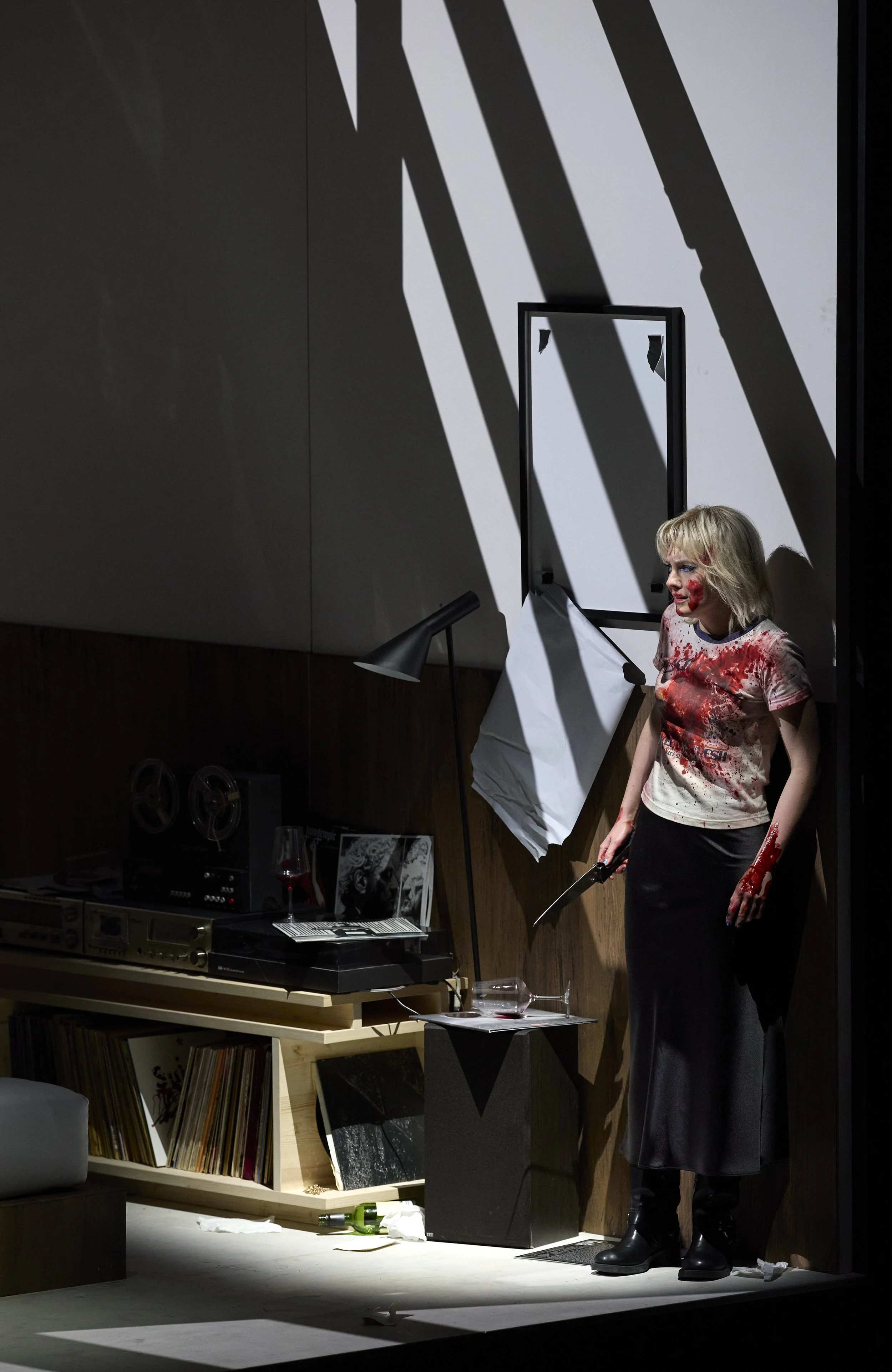 Frau steht in einem dunklen Raum, bekleidet mit blutigem T-Shirt und langem schwarzem Rock, mit einer funkelnden Klinge in der Hand. Sie ist blutverschmiert, hat blutverschmierte Haare und steht vor einem Spiegel, der von Lichtstrahlen beleuchtet wird, während der Raum eine Vinylsammlung, Bücher und eine Weinflasche enthält.