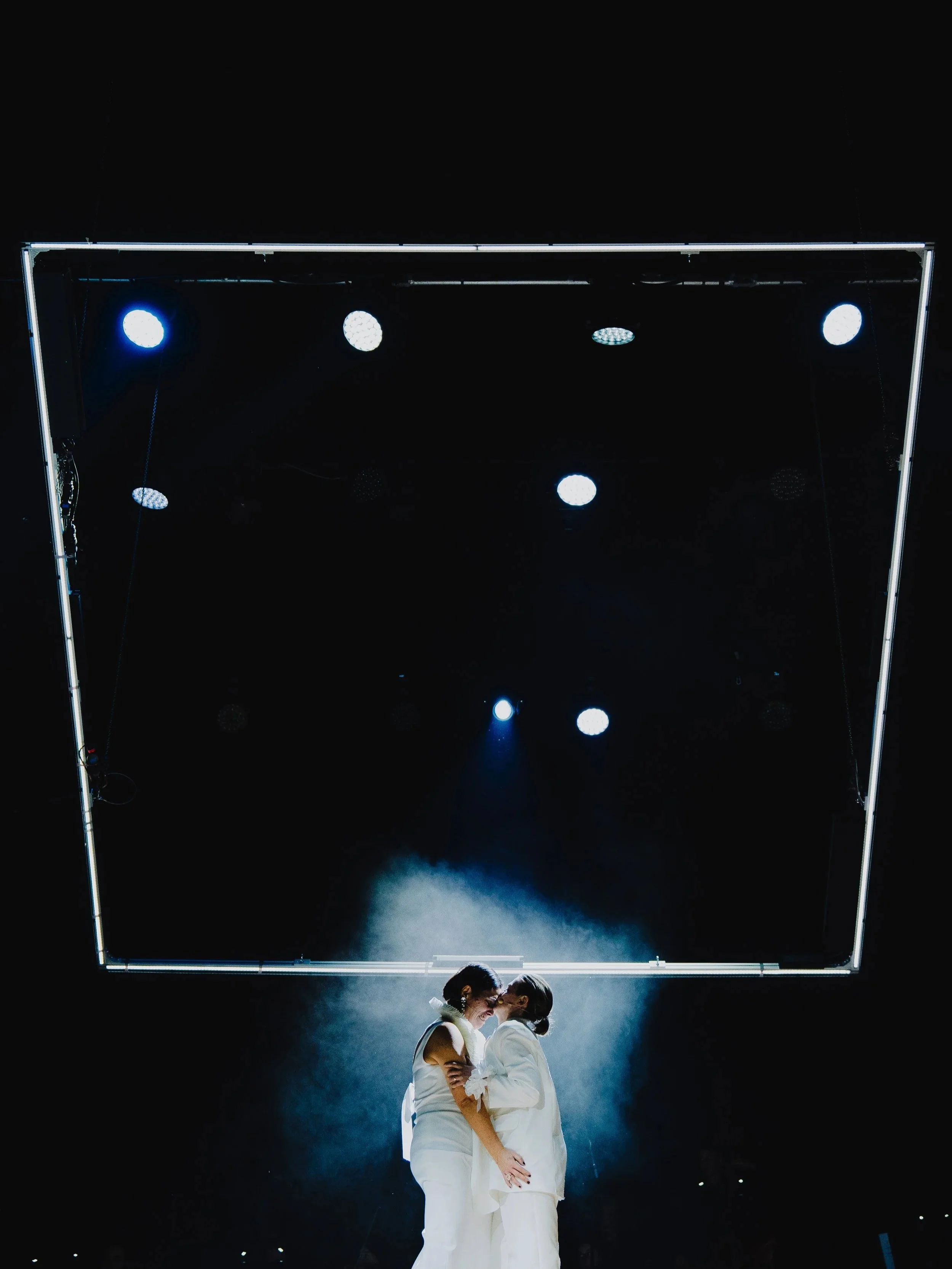 Zwei Frauen in weißem Outfit stehen sich berührend unter einer Bühnenbeleuchtung, schwarzer Hintergrund, modernes Bühnen-Setup.