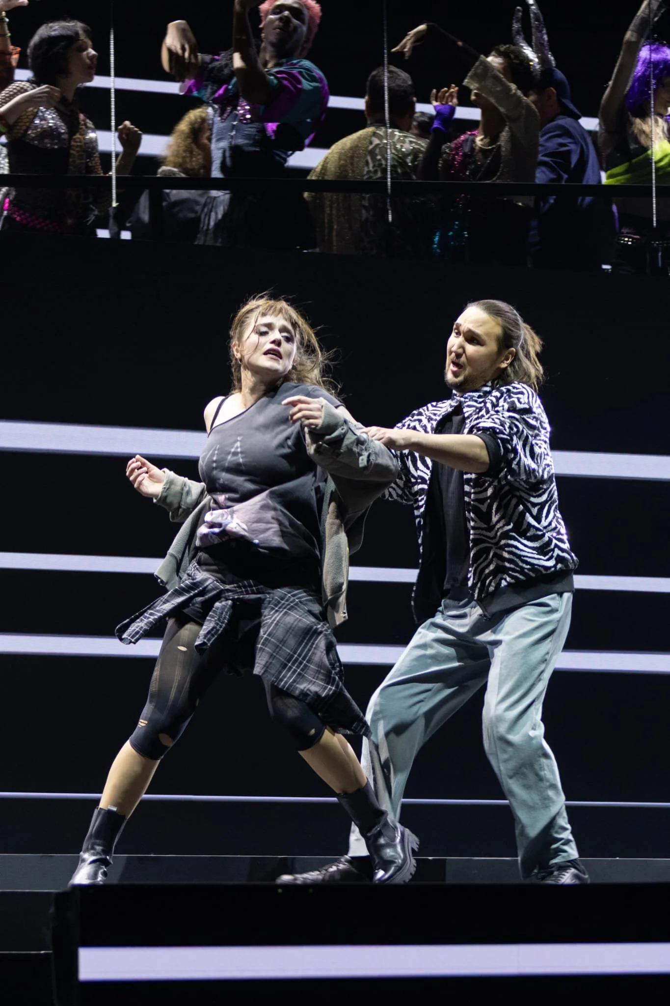 Zwei Schauspieler in kontroverser Szene auf einer Bühne, umrahmt von mehreren Zuschauern im Hintergrund, in einem Theaterstück.