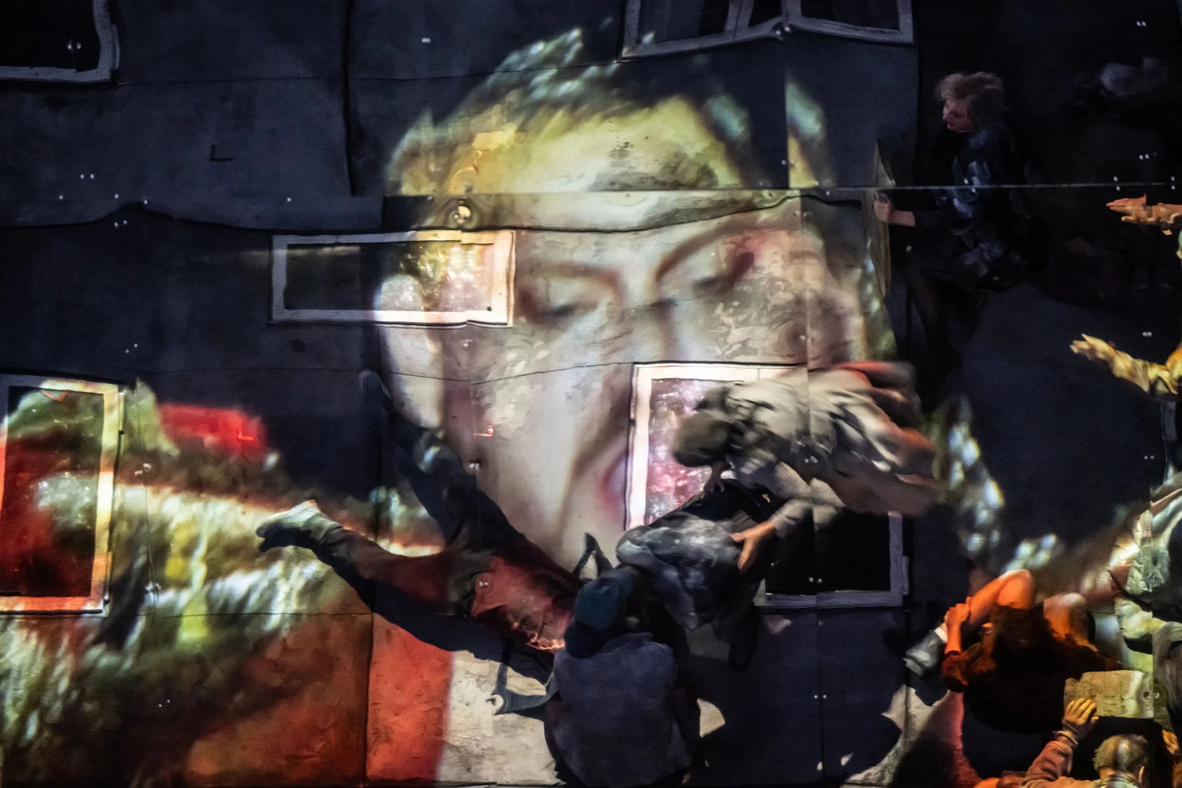 Menschen schauen auf eine große, projizierte Darstellung eines Kopfes, wahrscheinlich eines Künstlers, auf eine Wand, während sie den Bildschirm betrachten.