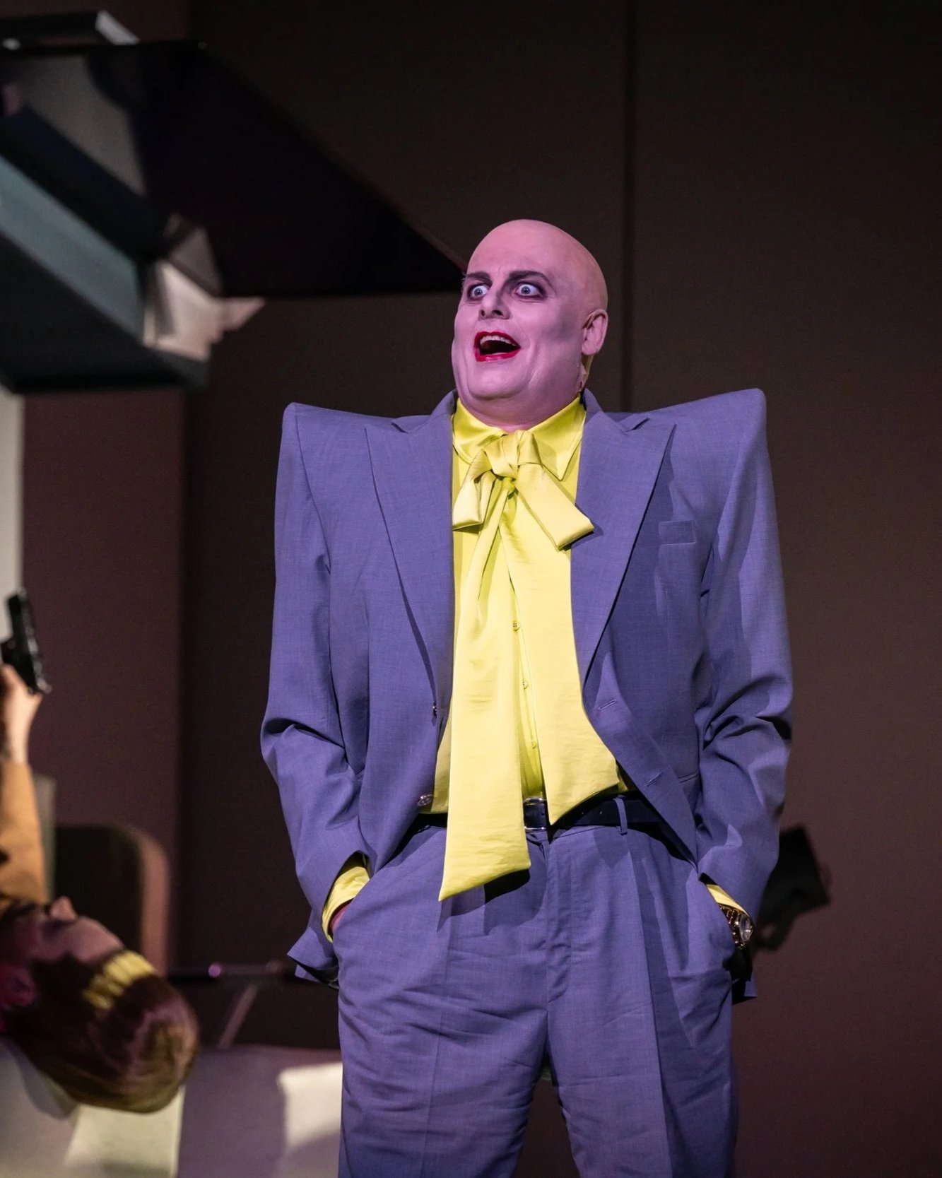 Eine Person in einem grauen Anzug mit einer gelben Bluse und Fliege steht vor einer Kamera. Die Person sieht überrascht oder schockiert aus.