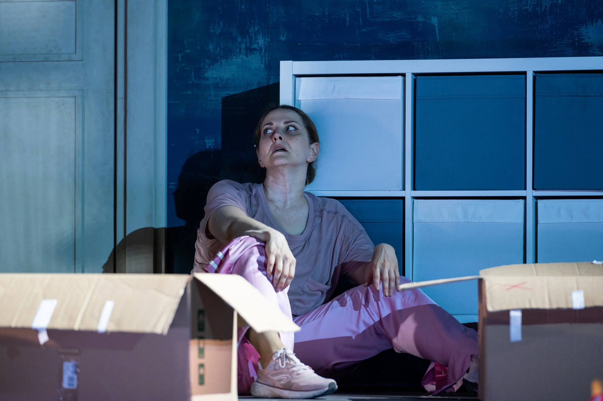 Eine erschöpfte Frau sitzt auf dem Boden zwischen offenen Kartons in einem dunklen Raum, mit einem verzerrten Ausdruck auf dem Gesicht.