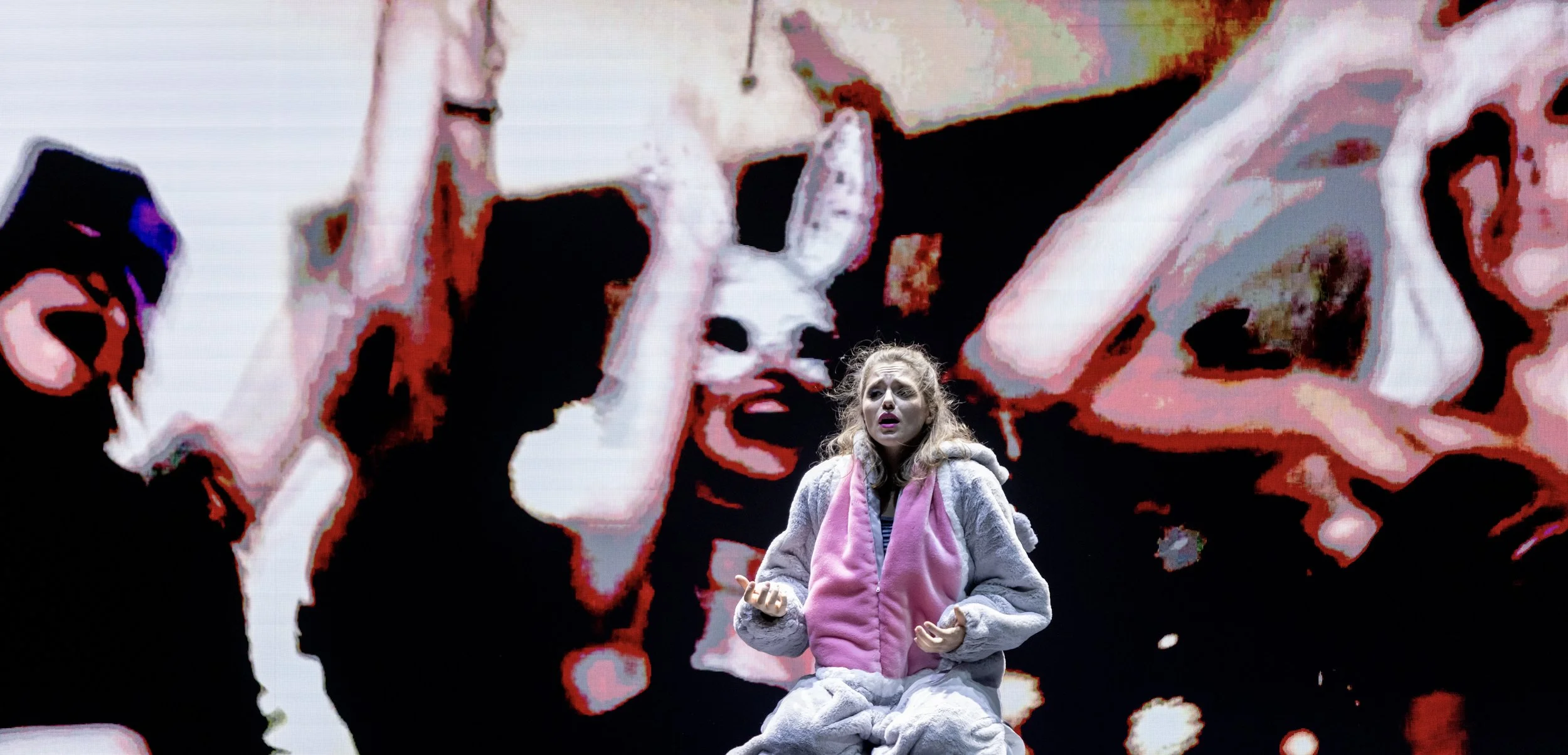 Eine junge Frau sitzt auf der Bühne mit einem Schauspiel im Hintergrund, das verzerrt wirkt mit Gesichtern und Händen. Sie trägt einen rosa und grauen Jacke und scheint in einer emotionalen Szene zu sein.