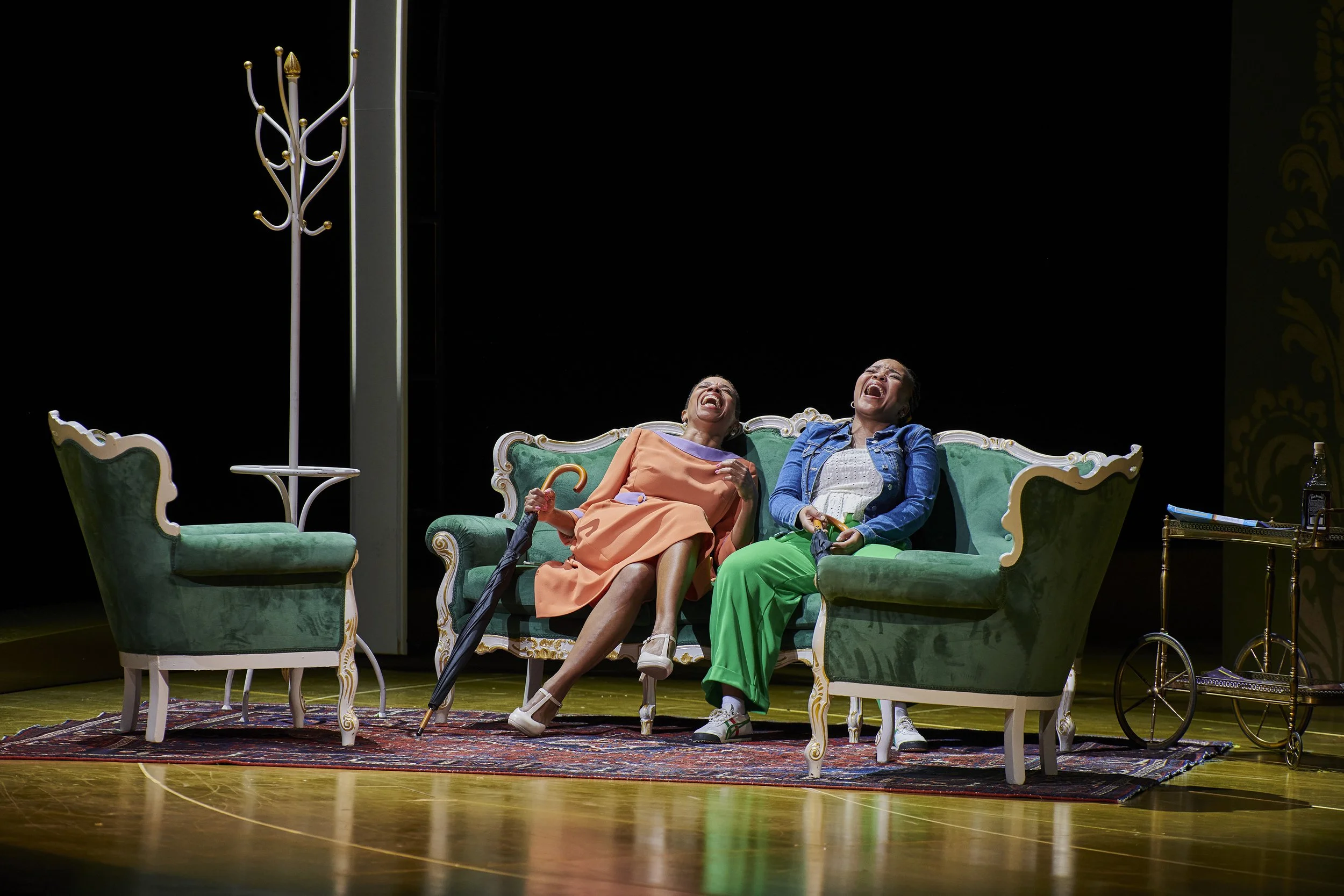 Zwei Frauen lachen auf einer grünen Couch mit goldverzierten Beinen auf einer Bühne, umgeben von einem Teppich, einem Stehleuchter, einem Beistelltisch und einem Barwagen.
