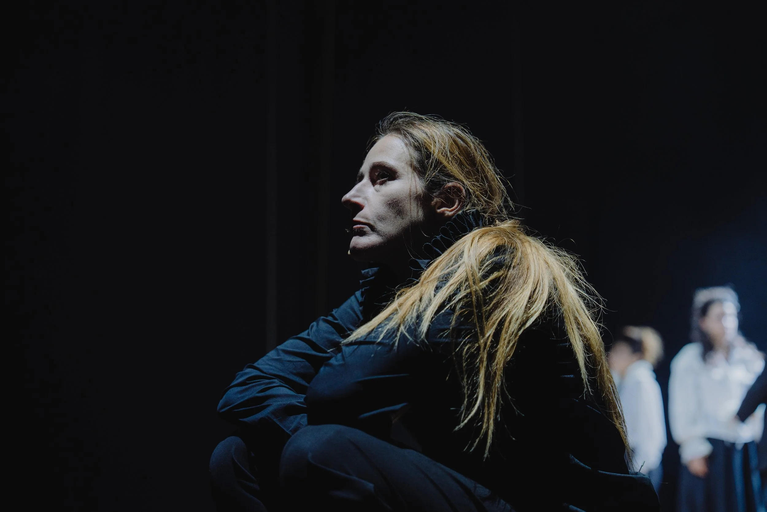 Frau mit langen, hellbraunen Haaren in dunkler Kleidung, in einer dunklen Umgebung mit mehreren Frauen im Hintergrund.