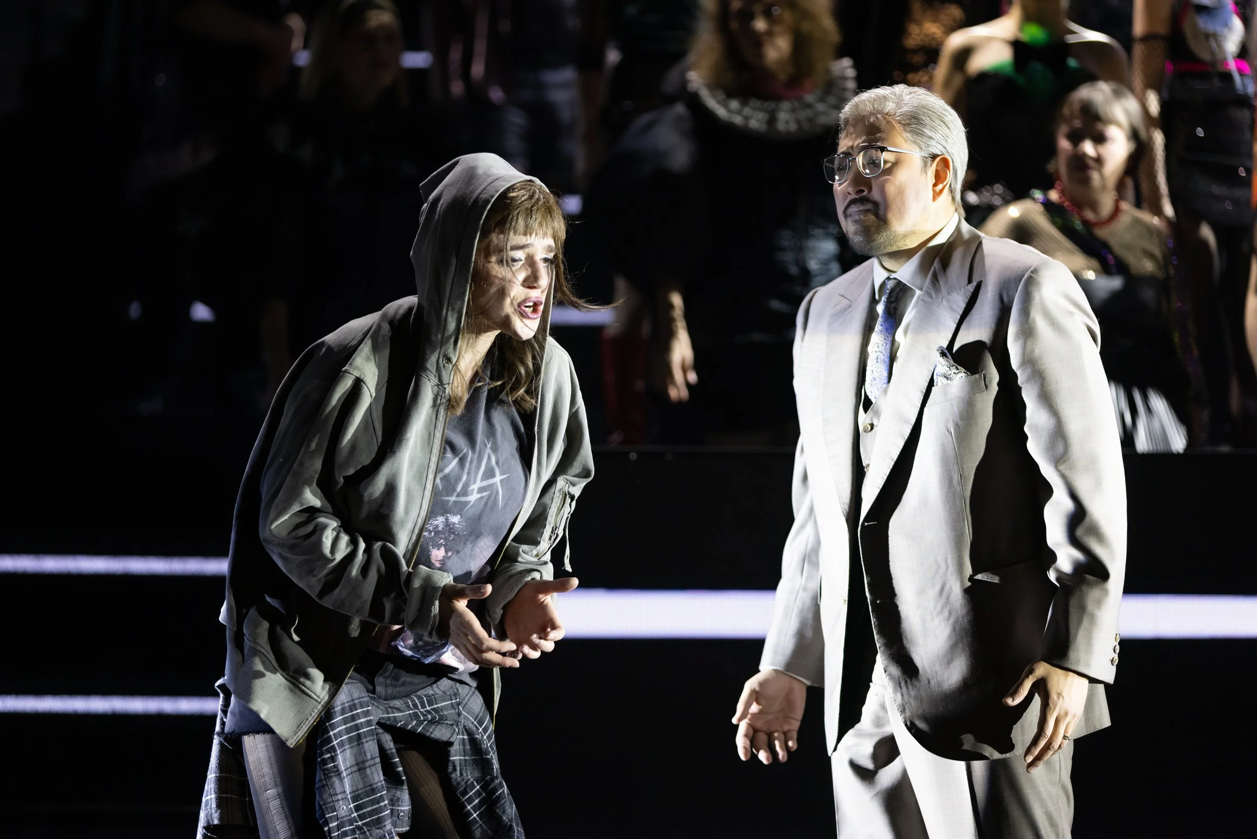 Zwei Menschen im Gespräch auf einer Bühne bei einer Theateraufführung. Der eine ist eine ältere Frau mit Brille, grauem Anzug und weißen Haaren, der andere eine jüngere Frau mit dunklem Haar in einem Hoodie, Jeansjacke und einem Kilt. Im Hintergrund 