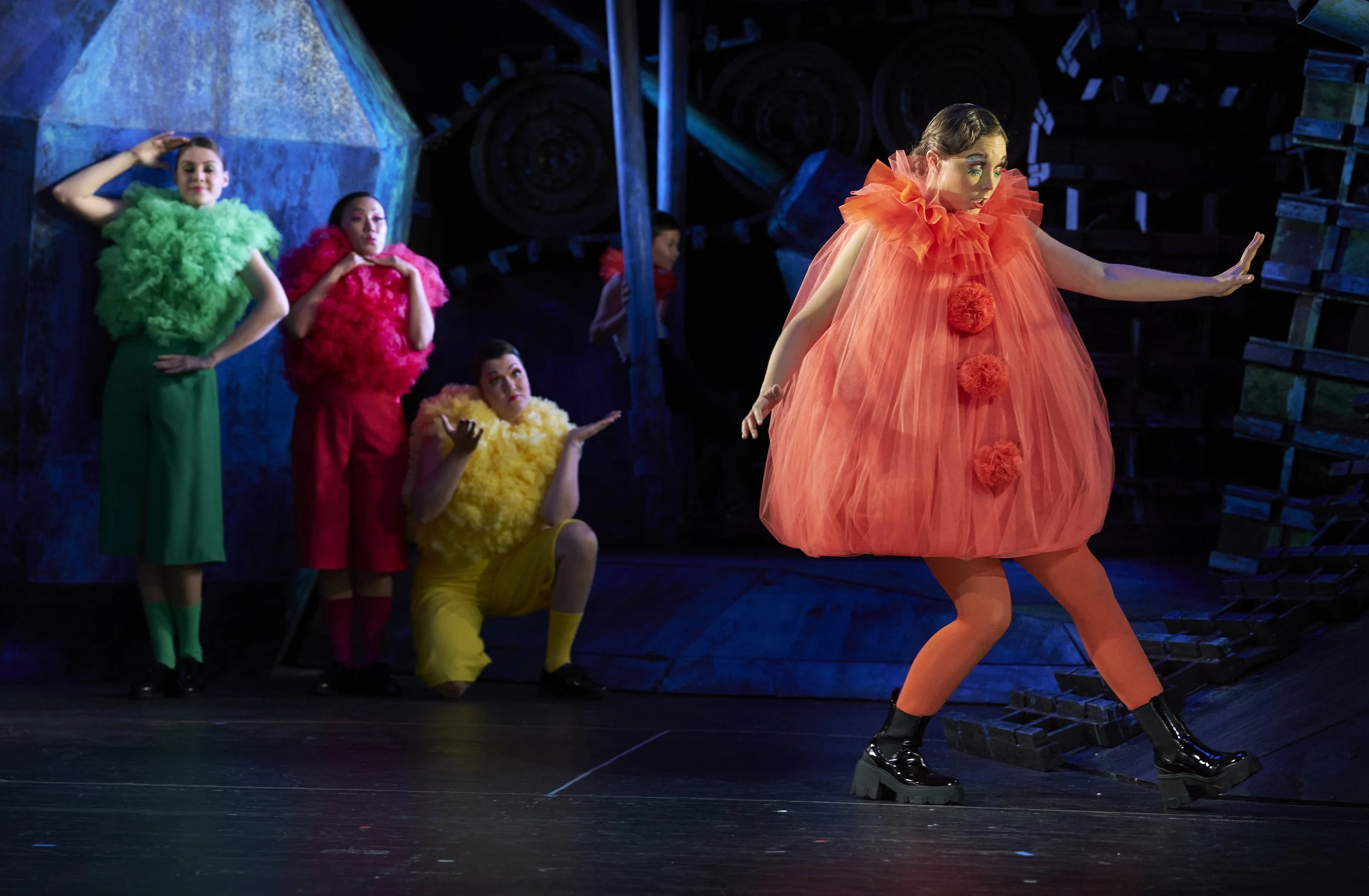 Eine Gruppe von vier Schauspielern in bunten Kostümen auf einer Bühne. Eine Frau in einem orangefarbenen Tüllkostüm und schwarzen Stiefeln posiert im Vordergrund, während die anderen drei hinter ihr in aufmerksamer Haltung stehen.