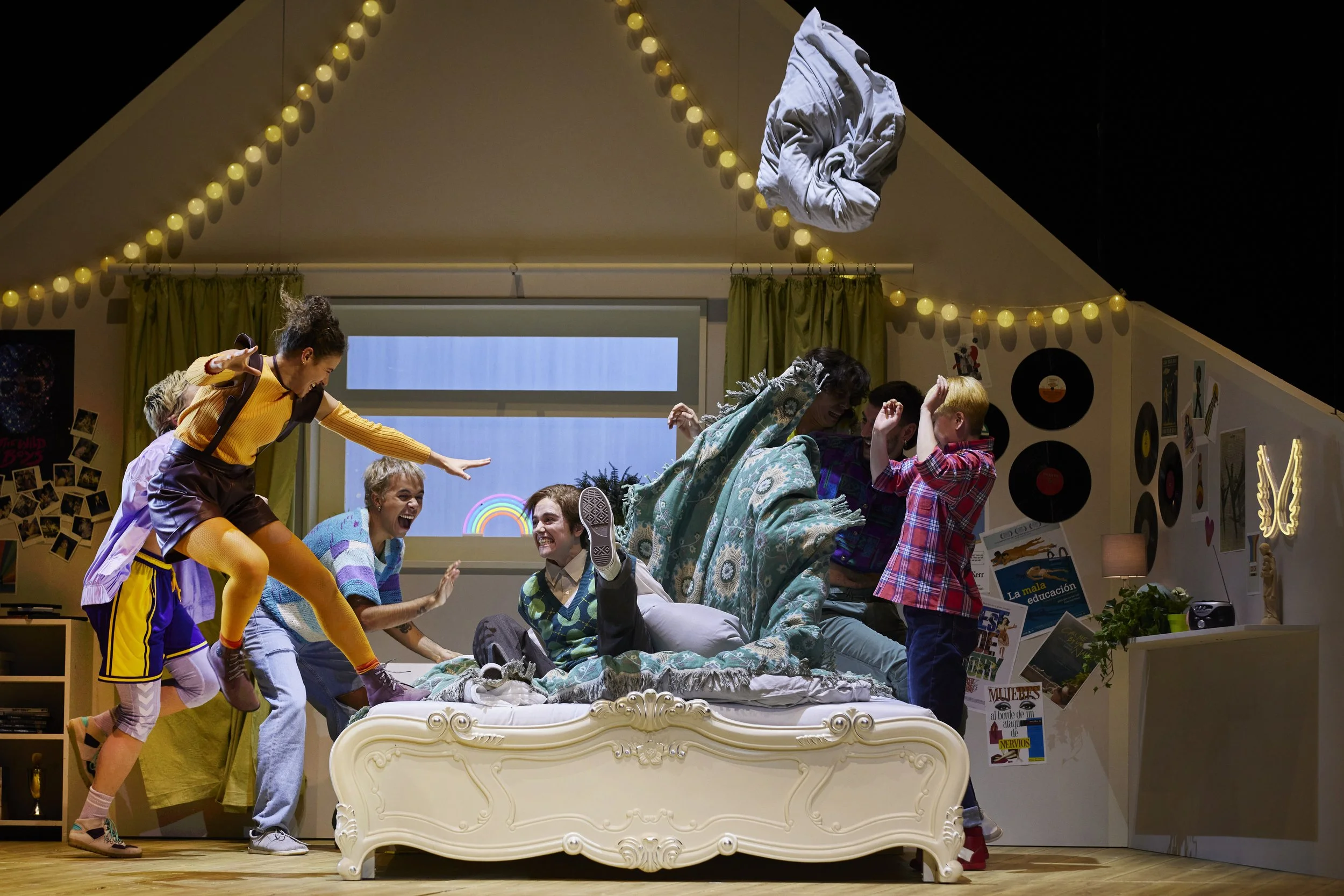 Szenenbild von einer Gruppe von jungen Menschen, die auf einem Bett in einem bunt dekorierten Zimmer spielen und lachen, mit Fenster im Hintergrund, Wanddekorationen und aufhängender Kleidung.