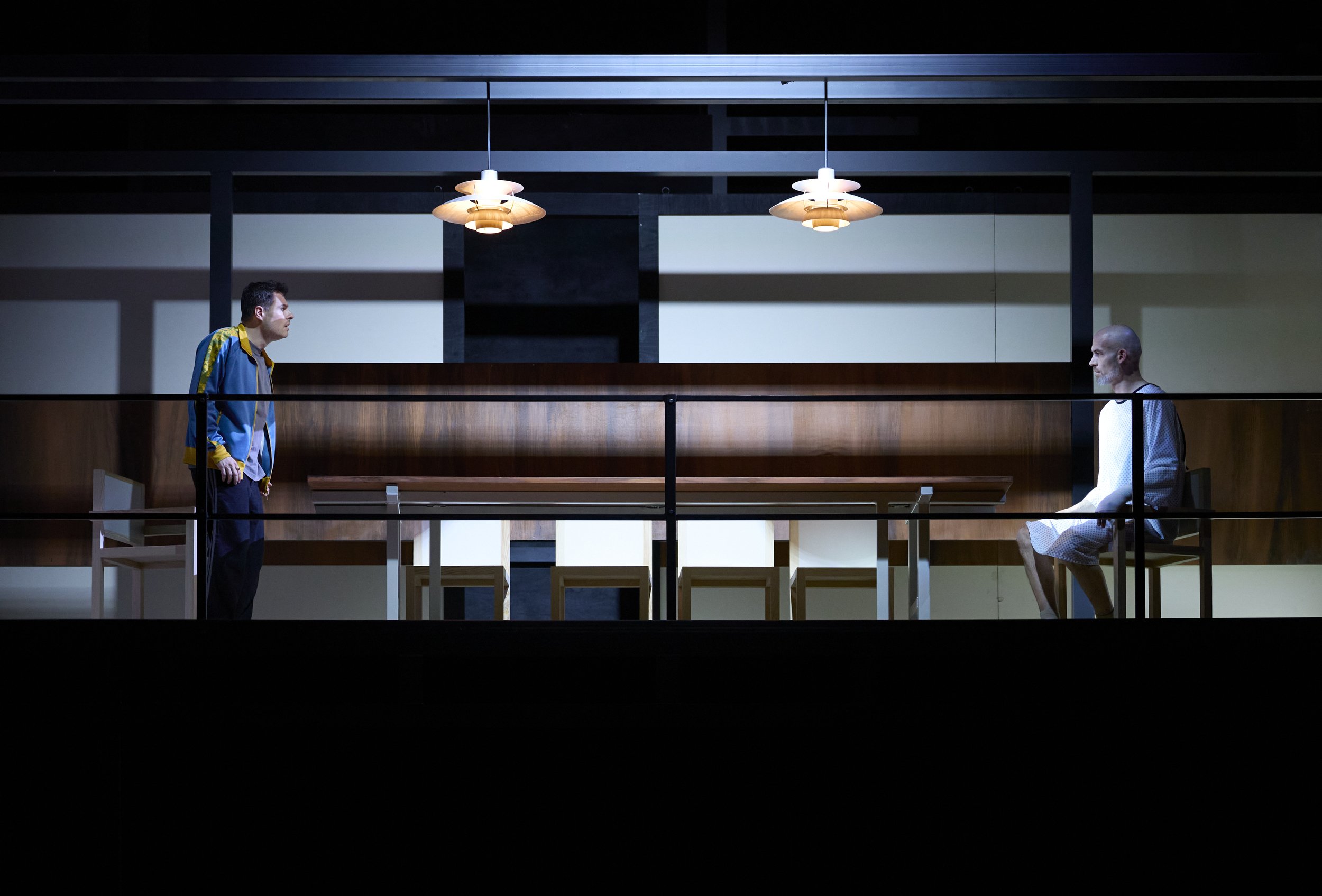 Zwei Männer sitzen in einem modernen Raum, gegenüber voneinander, mit Abstand. Sie schauen sich an, der eine trägt einen Pyjama, der andere sportliche Kleidung. Die Beleuchtung ist gedämpft, die Atmosphäre wirkt ernst.